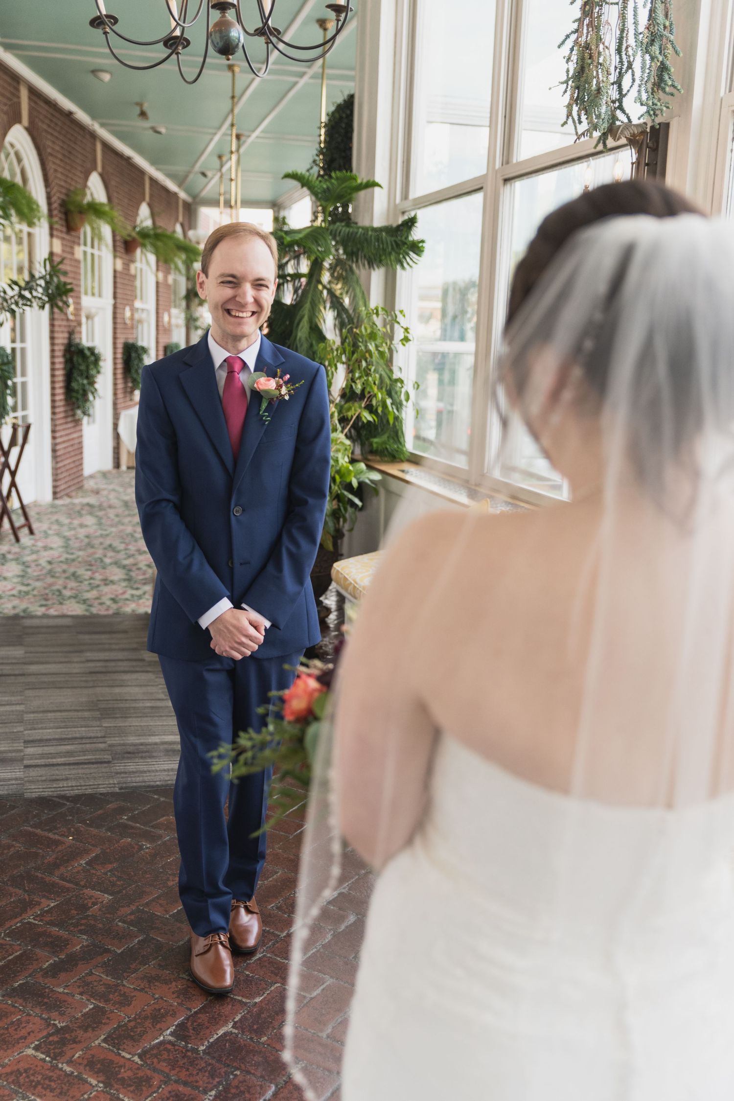 A wedding first look moment showing a groom in navy suit seeing his bride for the first time in a sunlit hallway.