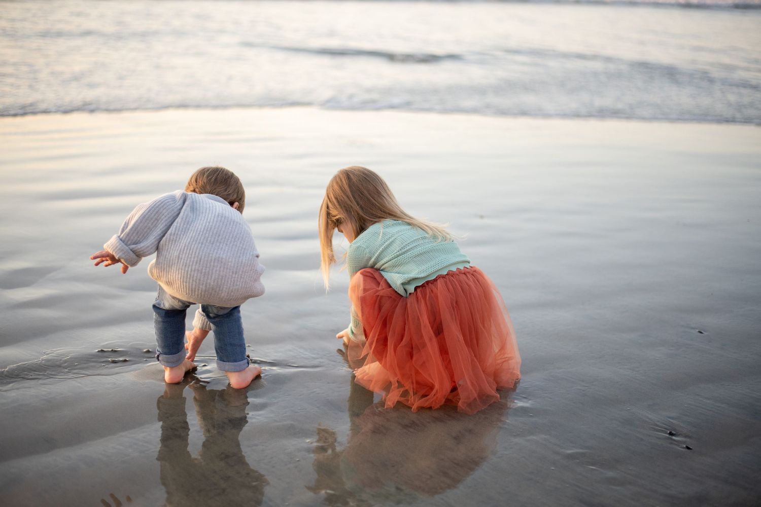 People exploring beach at sunset, bending down to examine shoreline.
