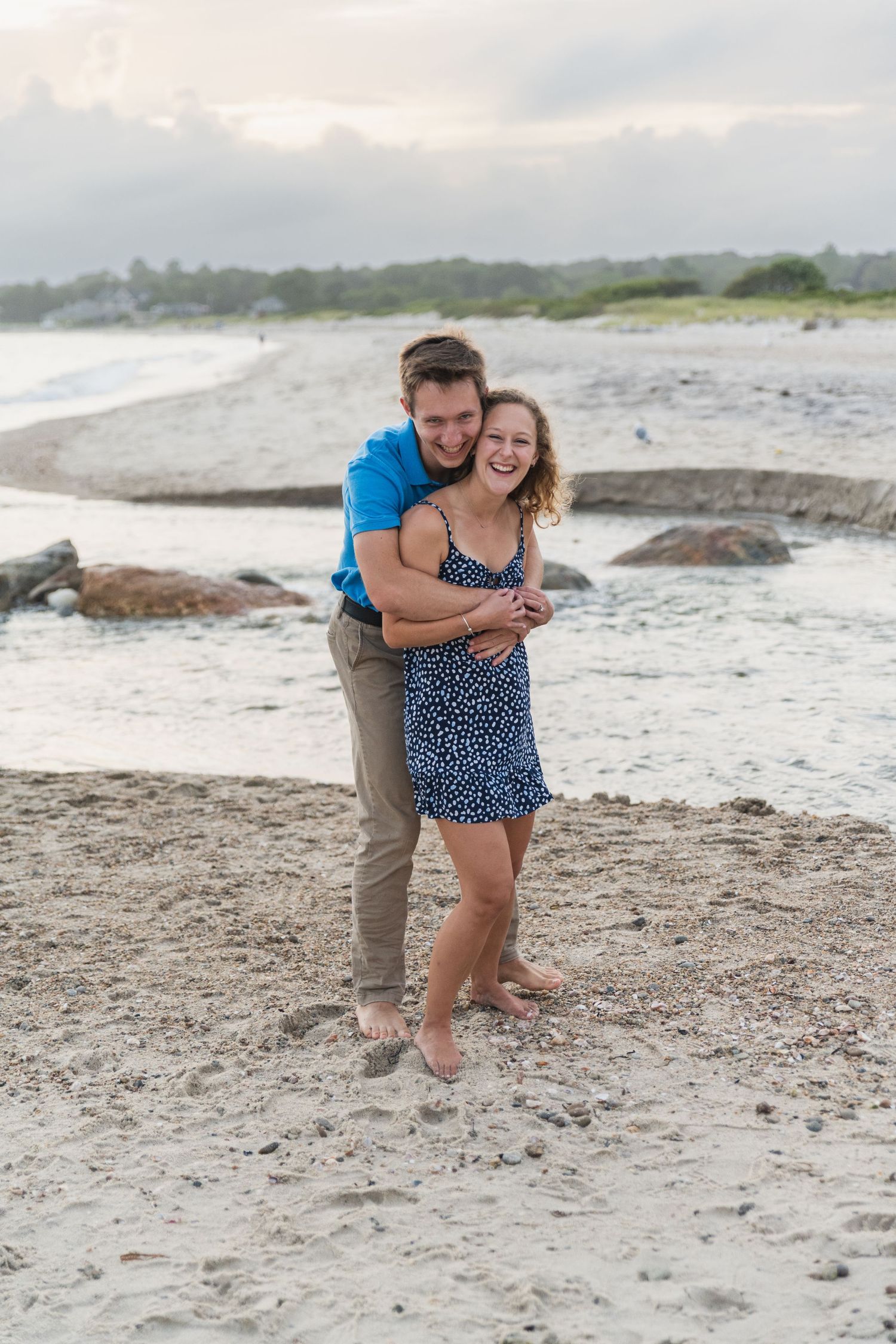 A joyful beach proposal at sunset shows two people embracing on the sand with waves crashing in the background.