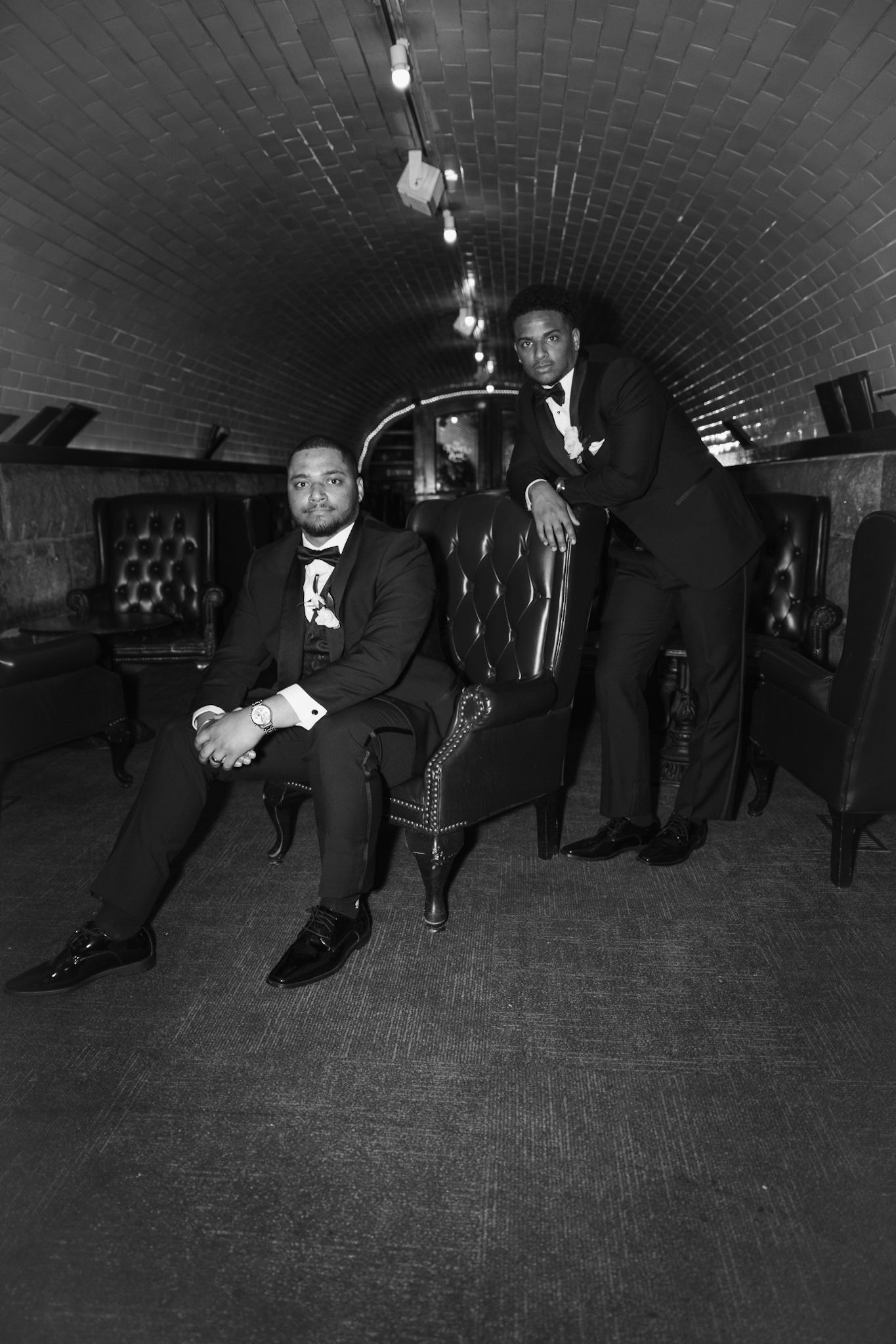 Group of groomsmen in formal black tuxedos posing together in arched brick hallway during wedding.