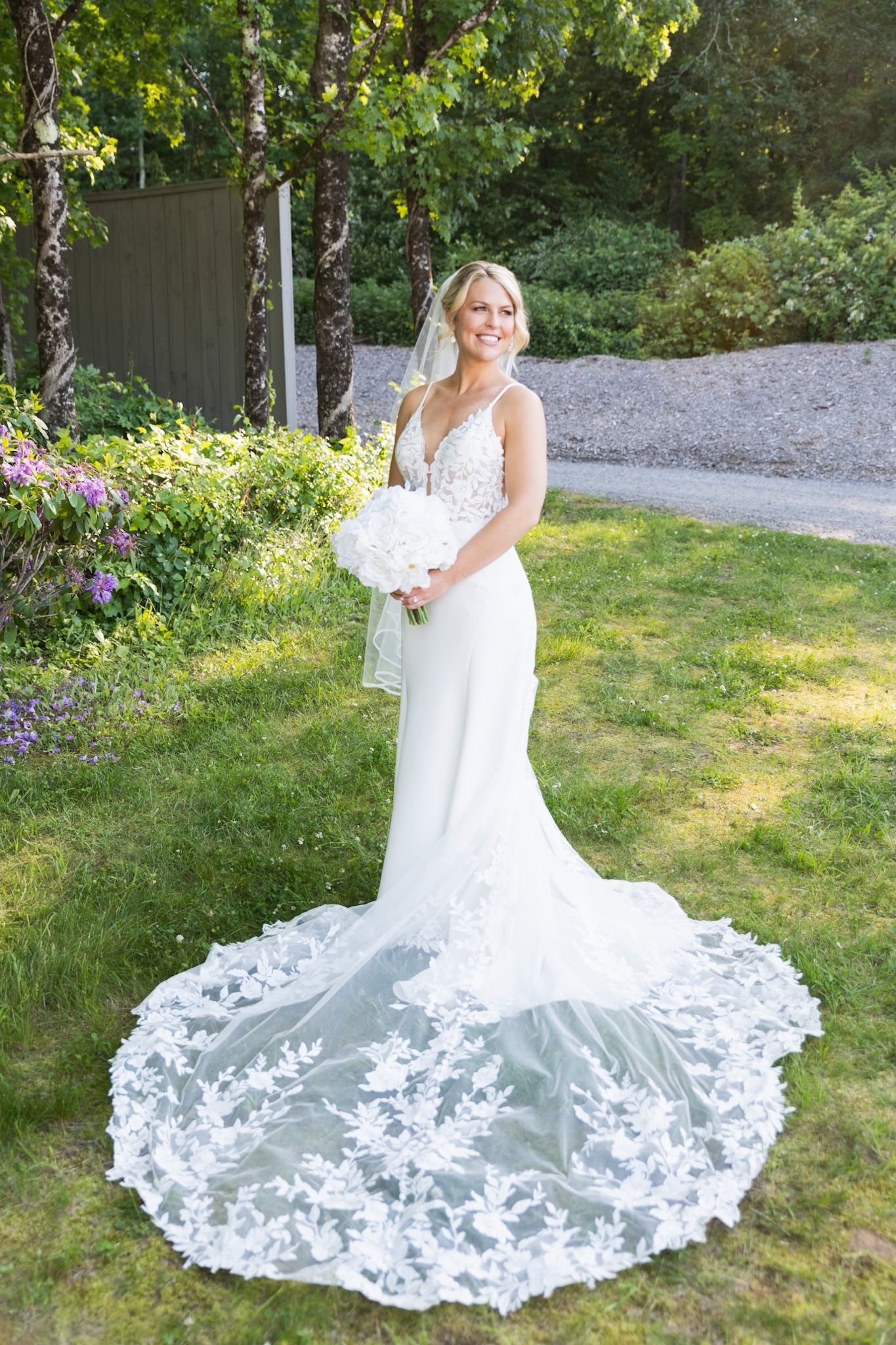 A wedding dress with an intricate lace train cascades across a garden lawn on a sunny day.