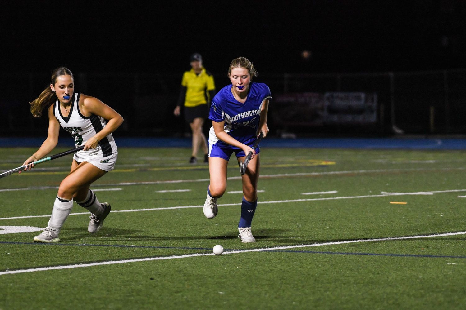 Two female field hockey players race for the ball during an evening match, one in blue and one in white.