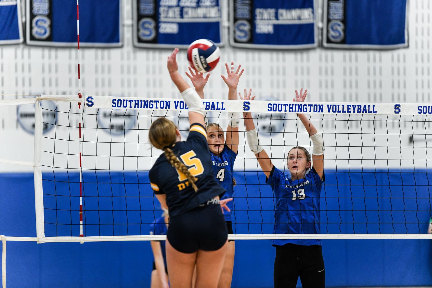 Southington volleyball players in blue uniforms block at the net during a competitive high school volleyball match.
