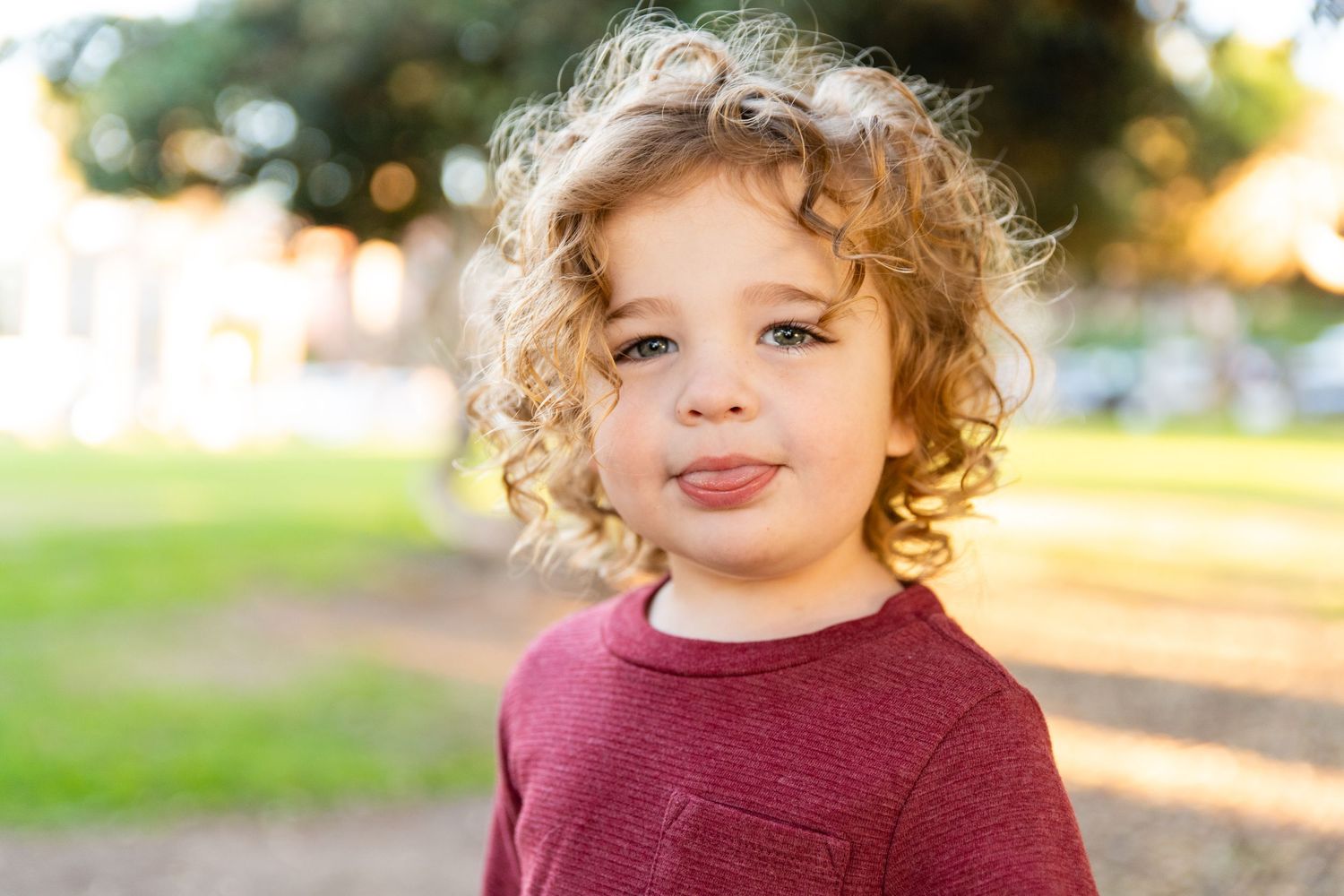 Child in burgundy sweater smiling outdoors with curly hair catching golden light.