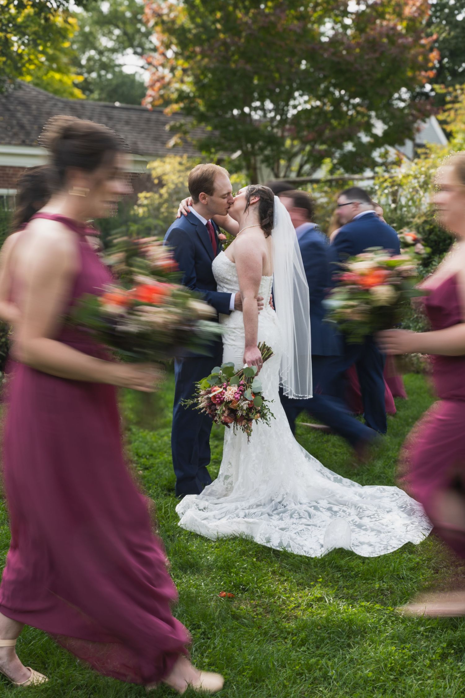 Wedding party in motion with bridesmaids in burgundy dresses blurred around couple sharing romantic moment on lawn.