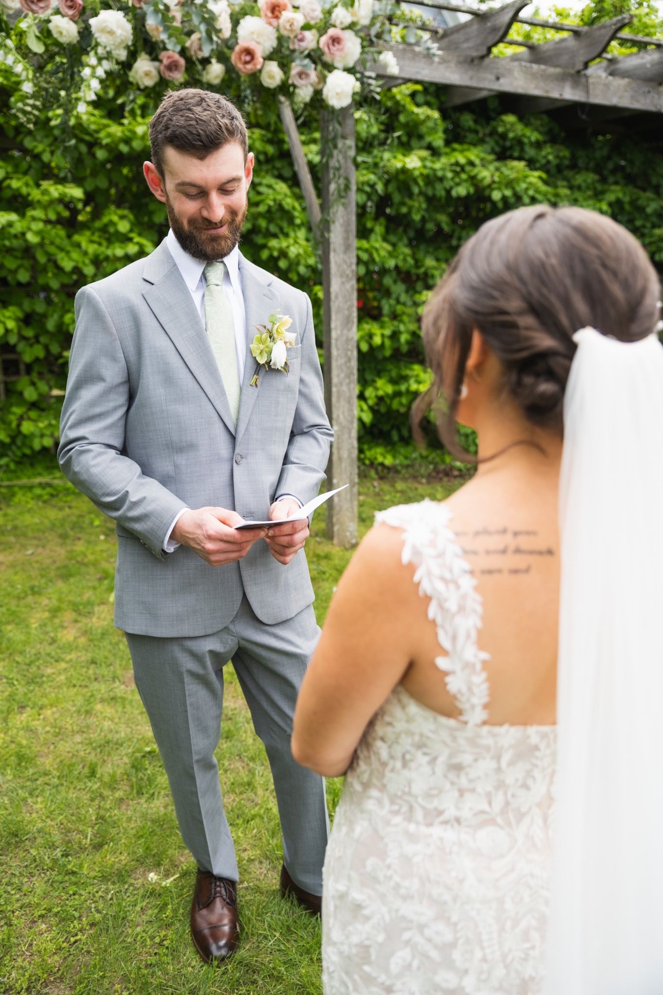 Groom in light gray suit reads vows under floral arch during outdoor ceremony.