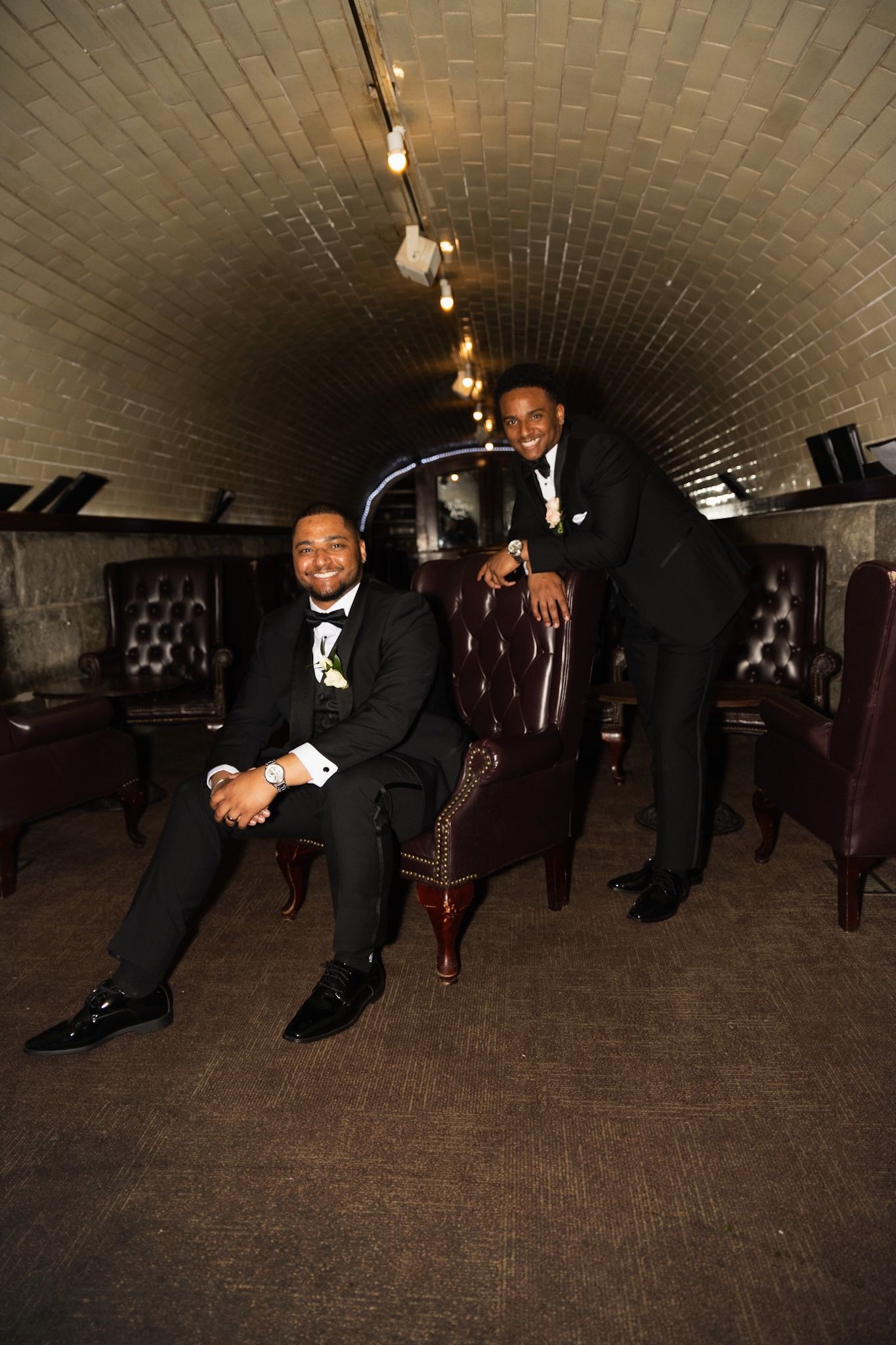 Two men in formal tuxedos posing in an elegant underground tunnel with arched brick ceiling.