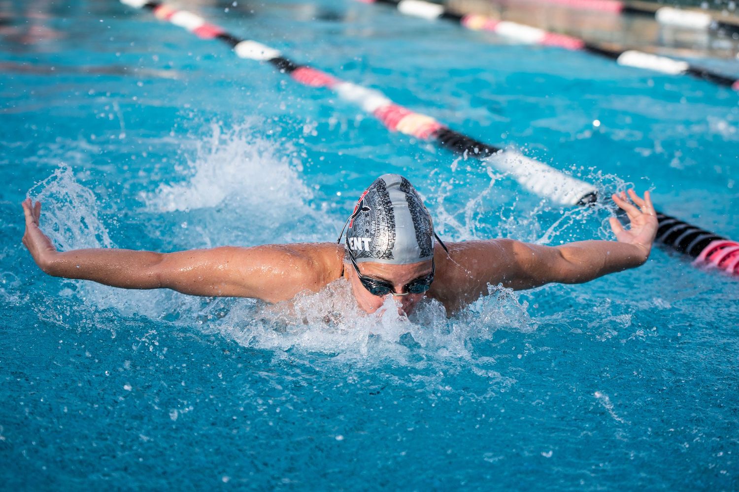 Competitive swimmer performing butterfly stroke in outdoor pool wearing goggles and swim cap during a race.