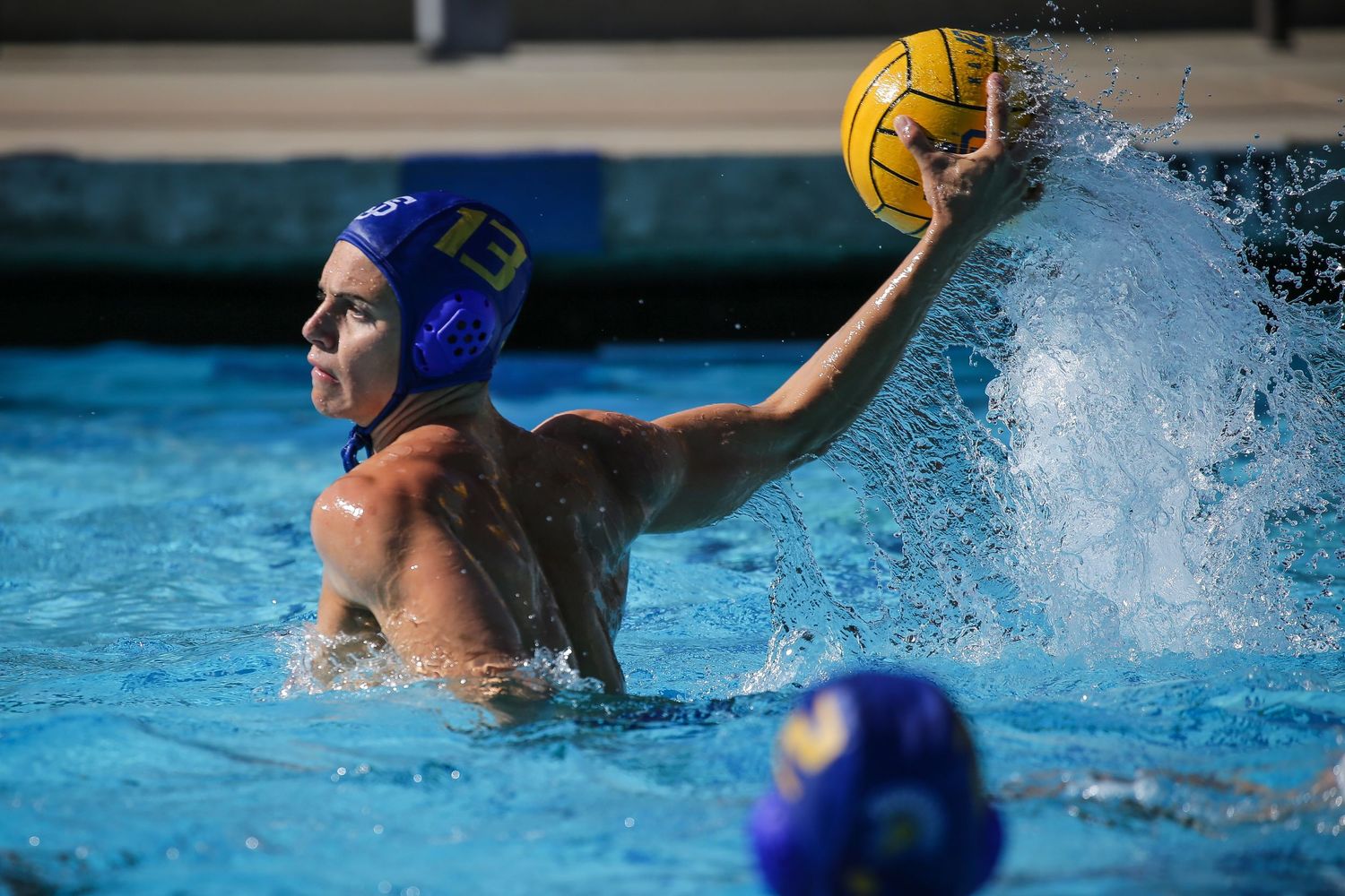 Water polo player in blue cap raises ball to throw during competitive pool match.