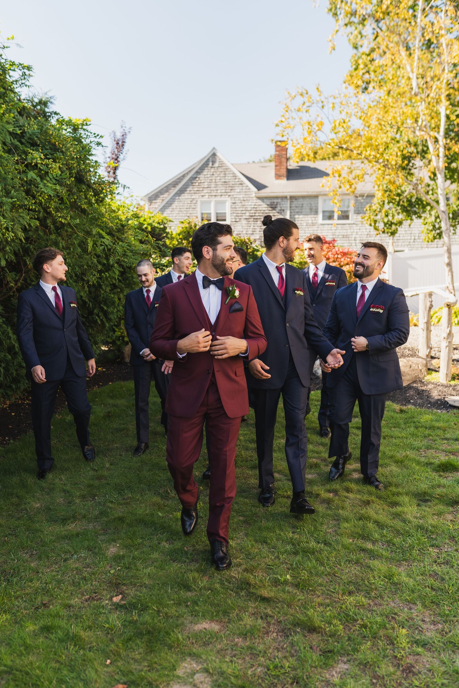 Groomsmen in burgundy and navy suits share laughter while standing on grass during outdoor wedding photos.