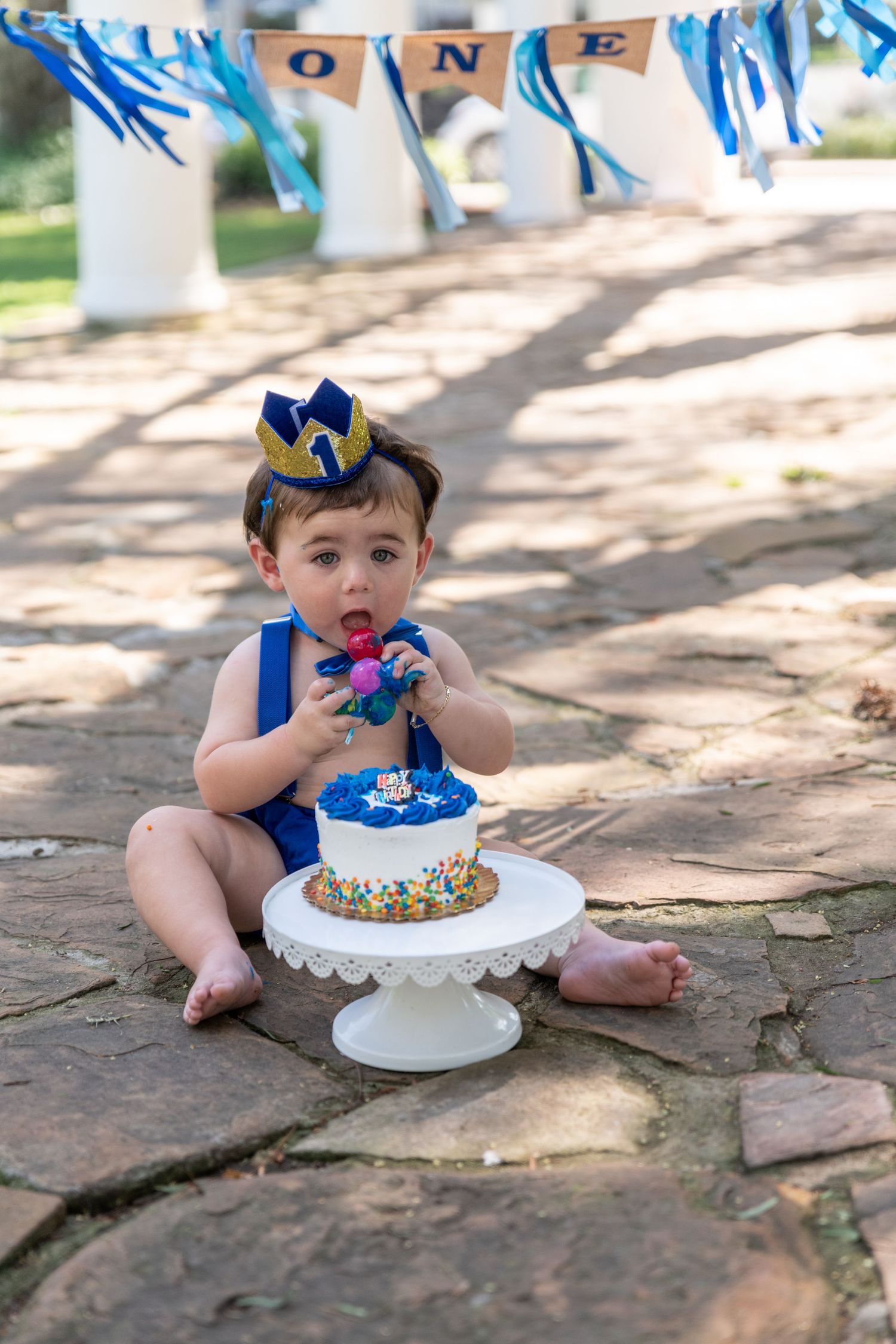 Baby in blue outfit and crown celebrates first birthday with cake outdoors.