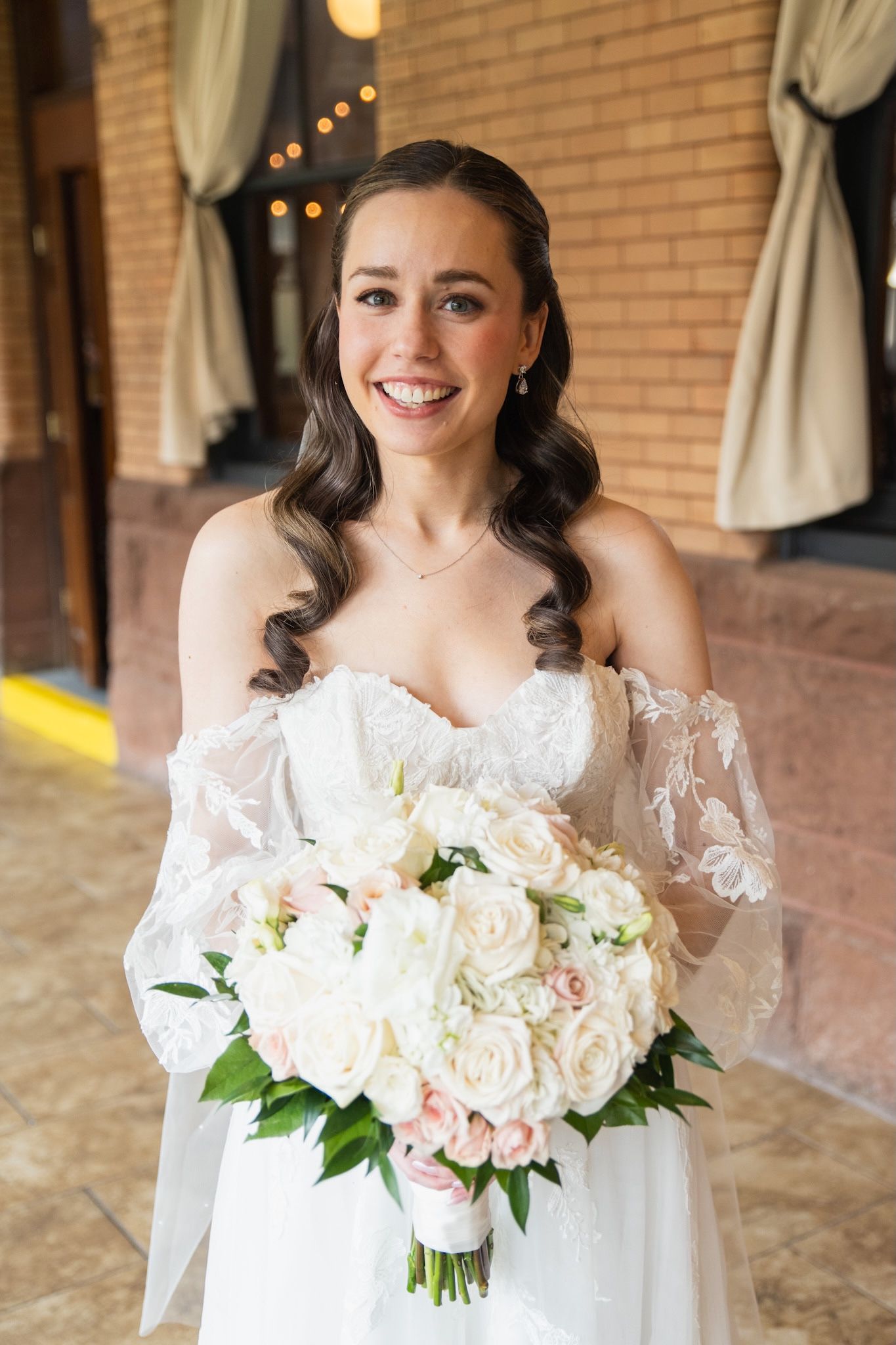 Bride holding elegant white and blush rose bouquet in off-shoulder lace wedding gown.