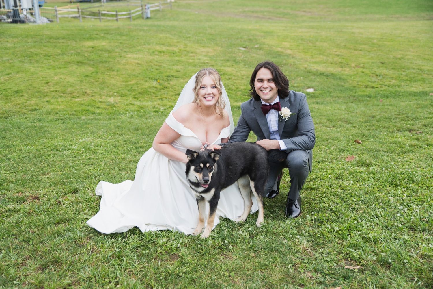 A wedding couple sitting on grass with their black and white dog.