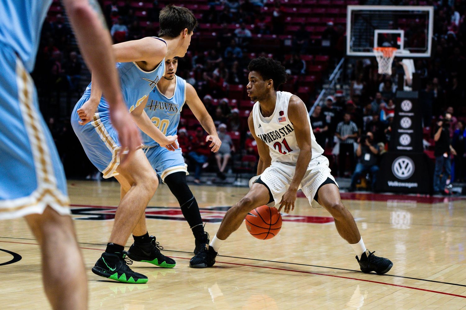 College basketball player in white jersey dribbles ball while defended by opponent in blue during indoor arena game.