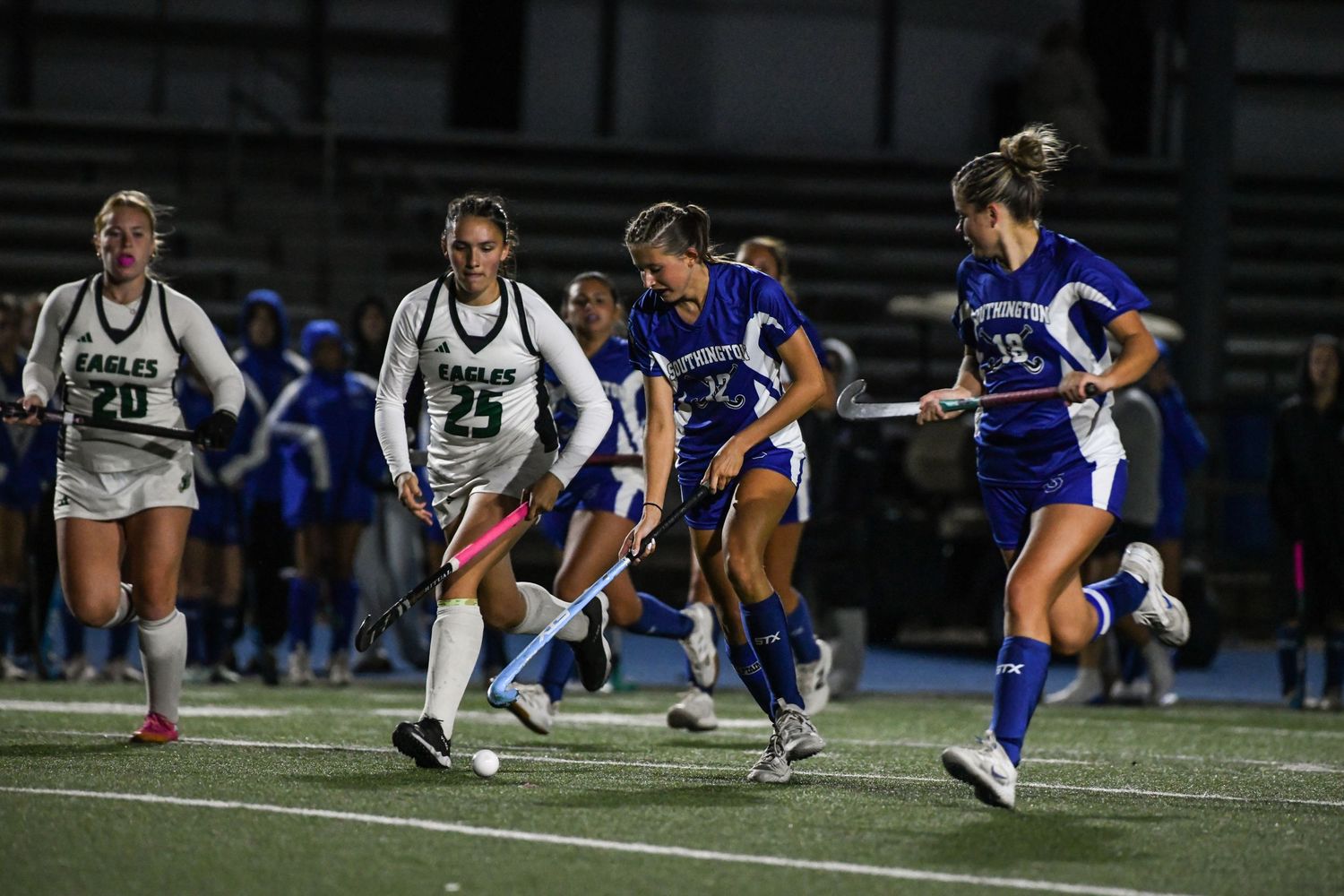 Female field hockey players in blue and white uniforms compete during a night game on a turf field.