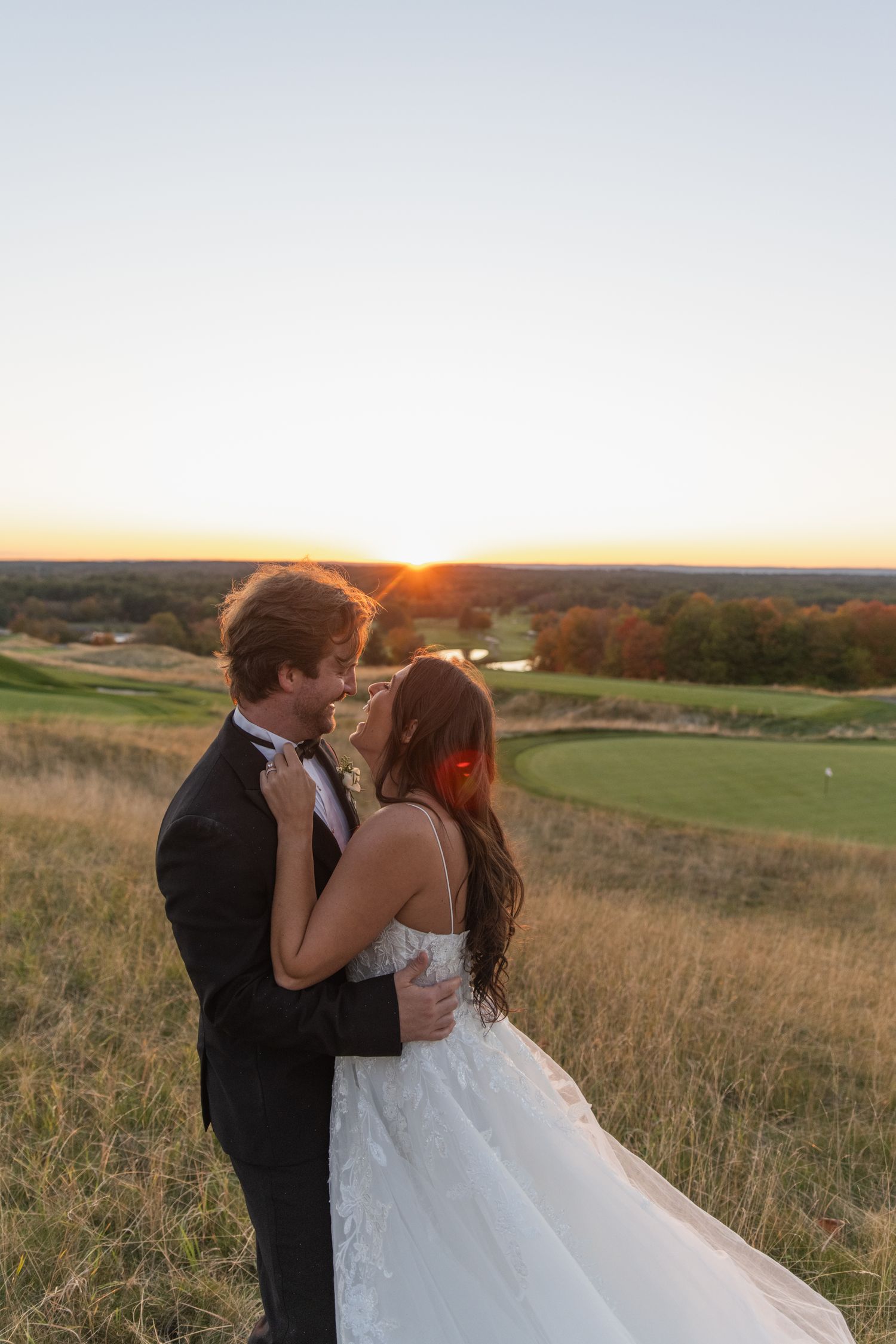 A couple in wedding attire embraces on a hilltop overlooking a golf course at sunset.