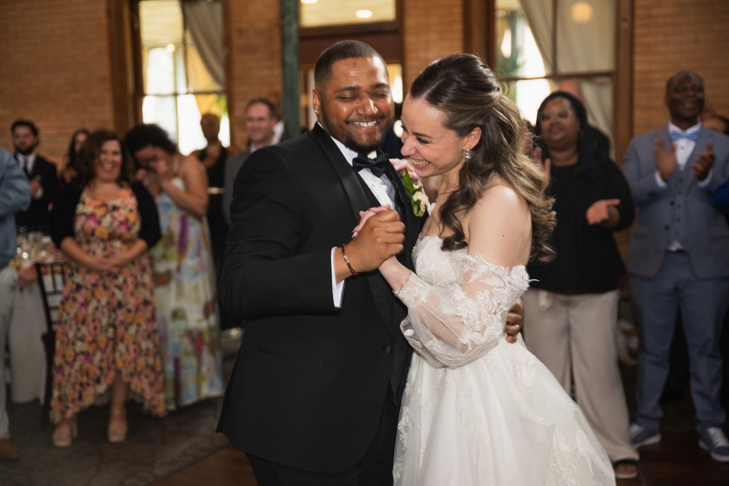 Newlyweds sharing their first dance surrounded by wedding guests in an indoor reception venue.