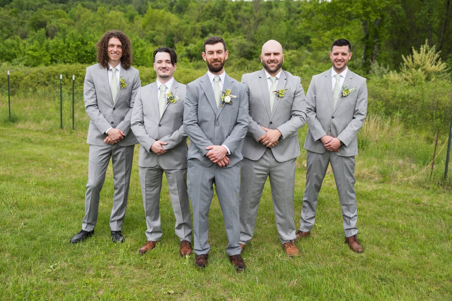 Groomsmen in matching light gray suits stand together on grass with boutonnieres.
