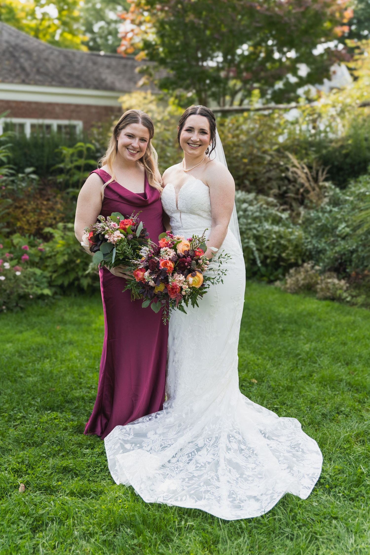 A wedding party member in burgundy and a bride in white lace hold vibrant fall-colored bouquets in a garden setting.