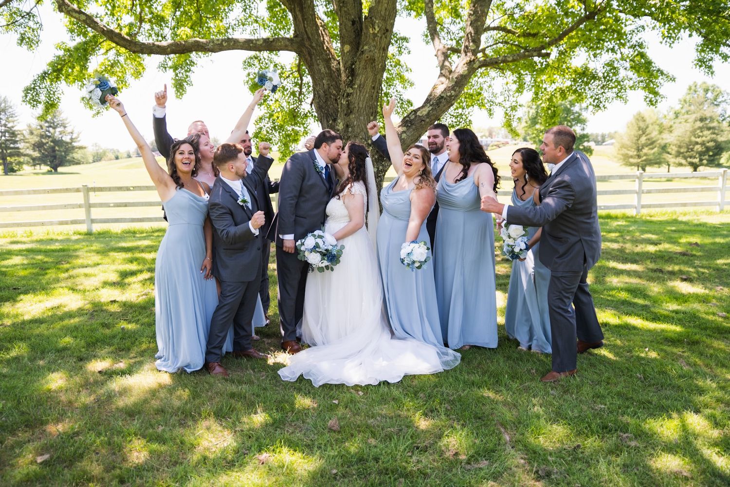 Wedding party poses joyfully under a tree in light blue and gray attire.