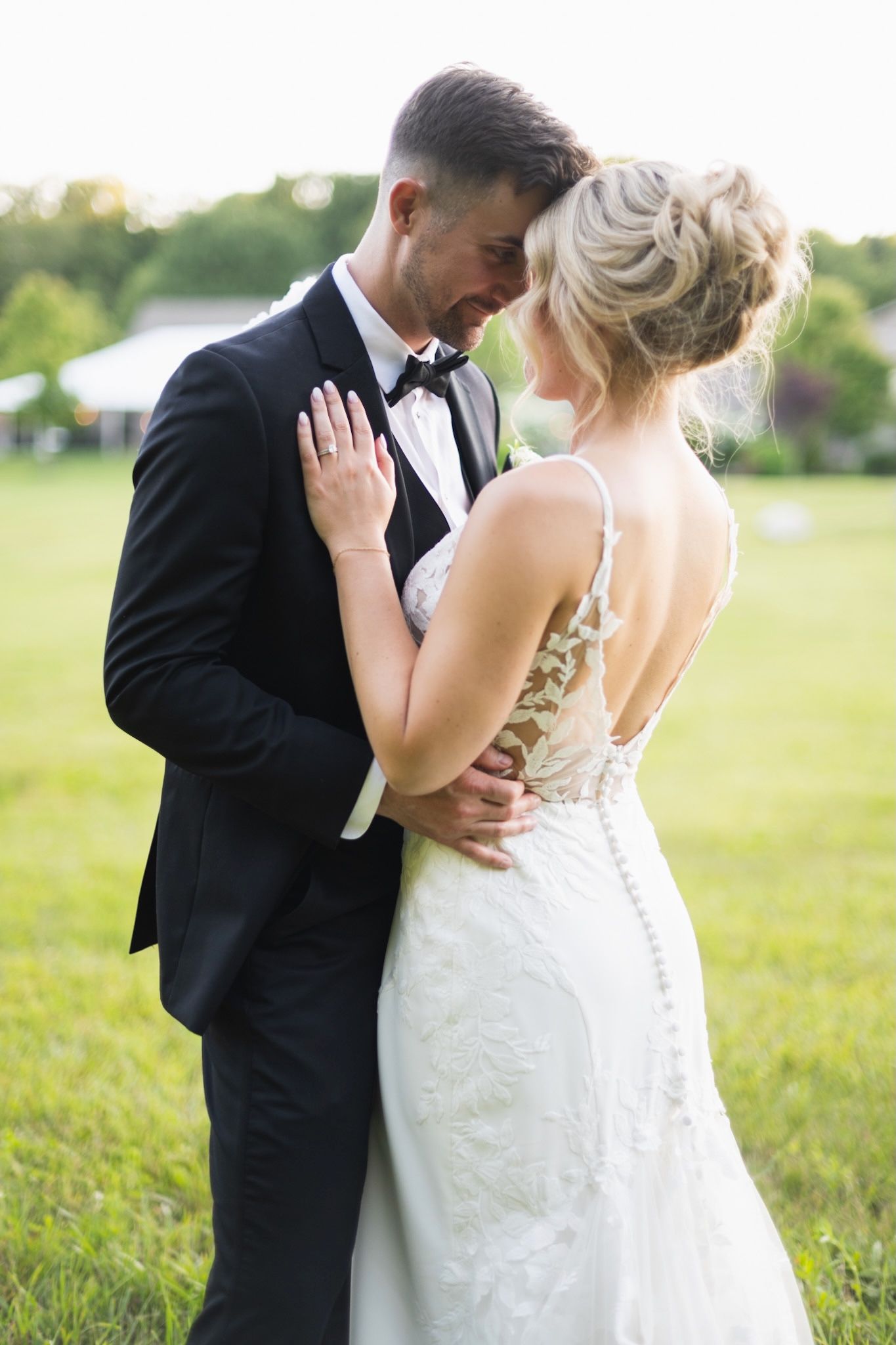 A romantic wedding portrait of a couple in formal attire standing in a sunlit field.