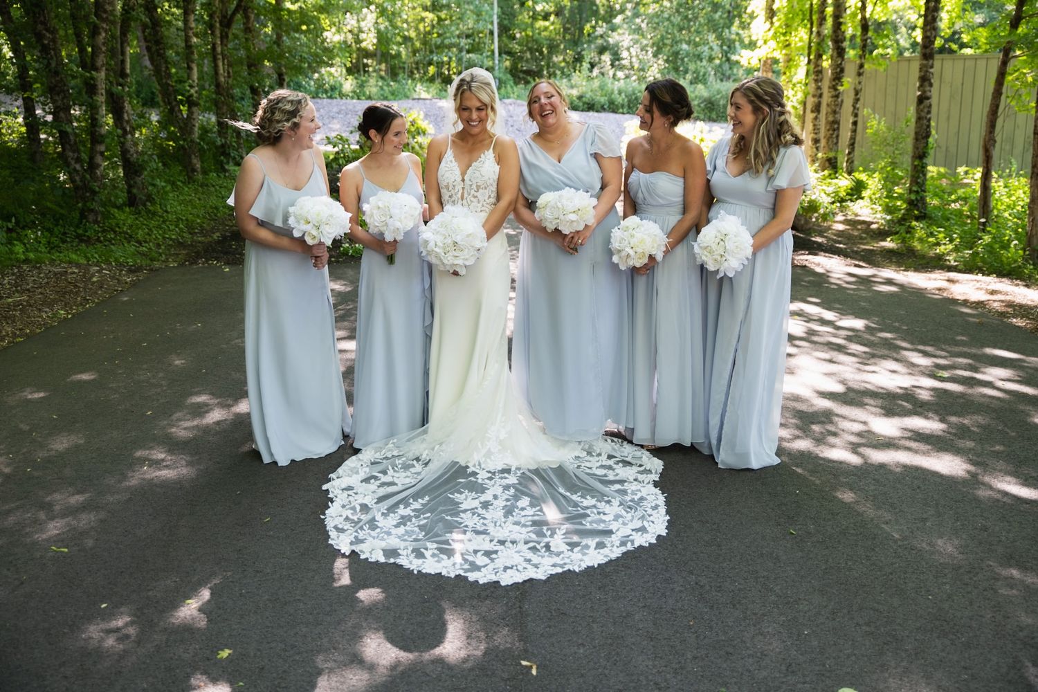 Bridesmaids in light blue dresses gather around bride with lace train on outdoor path.