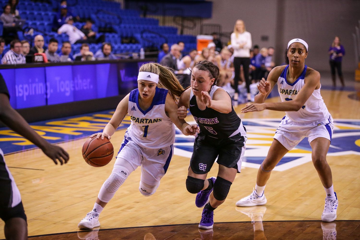 Women's college basketball players compete for the ball during an intense Spartans home game on a blue court.
