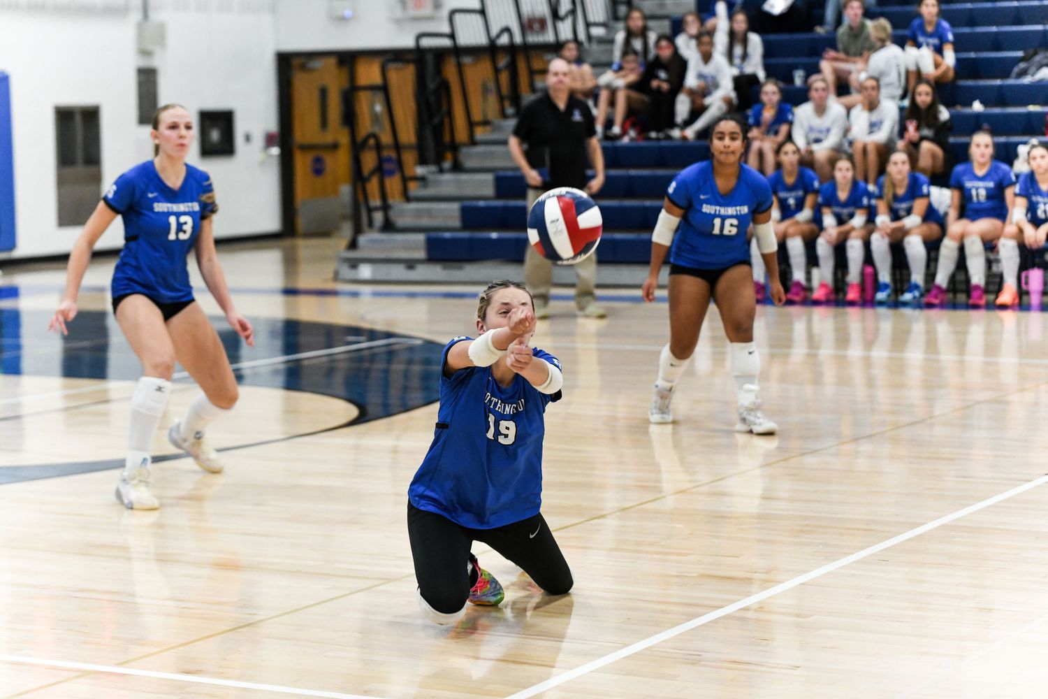 Female volleyball player in blue uniform digs a ball during an indoor match with teammates visible in background.