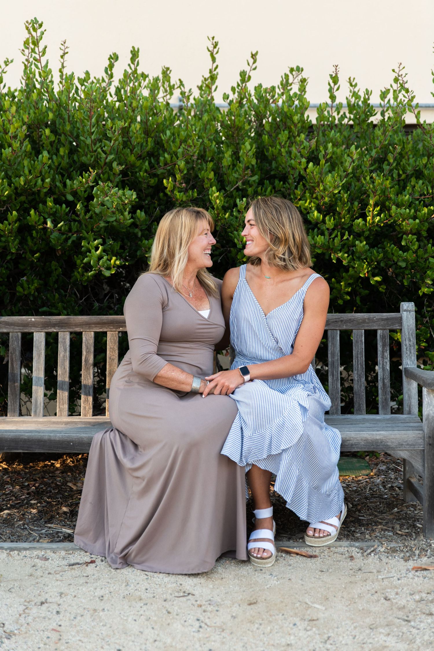 Two friends sharing laughter while sitting together on wooden bench by hedge.