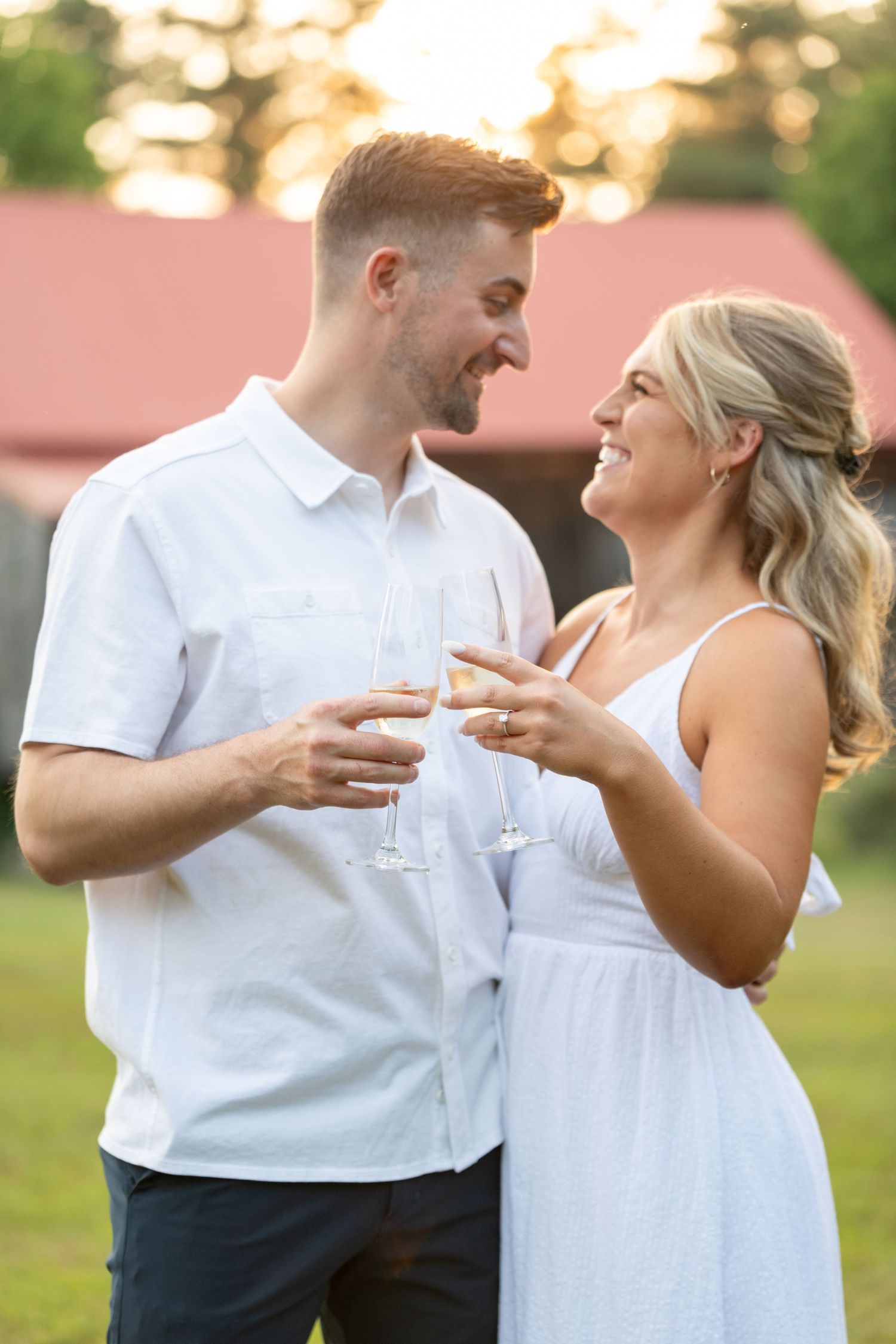Couple in white outfits sharing champagne and laughing during sunset photo session.