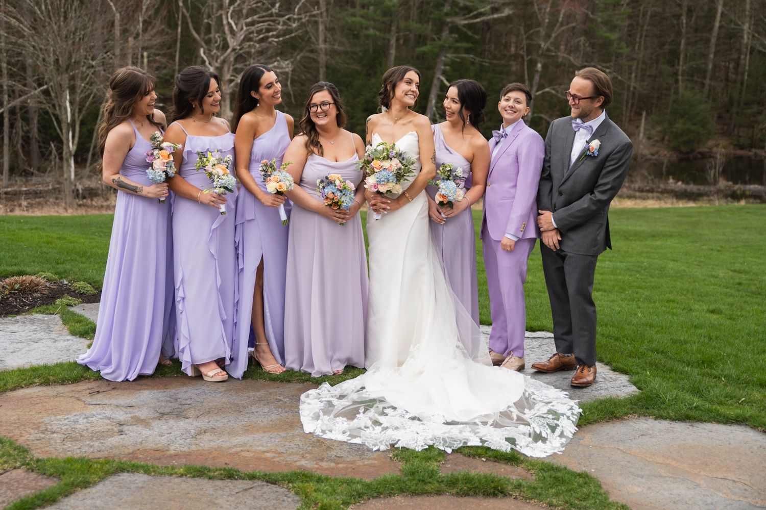 Wedding party in purple bridesmaid dresses and formal attire poses on lawn for group photo.