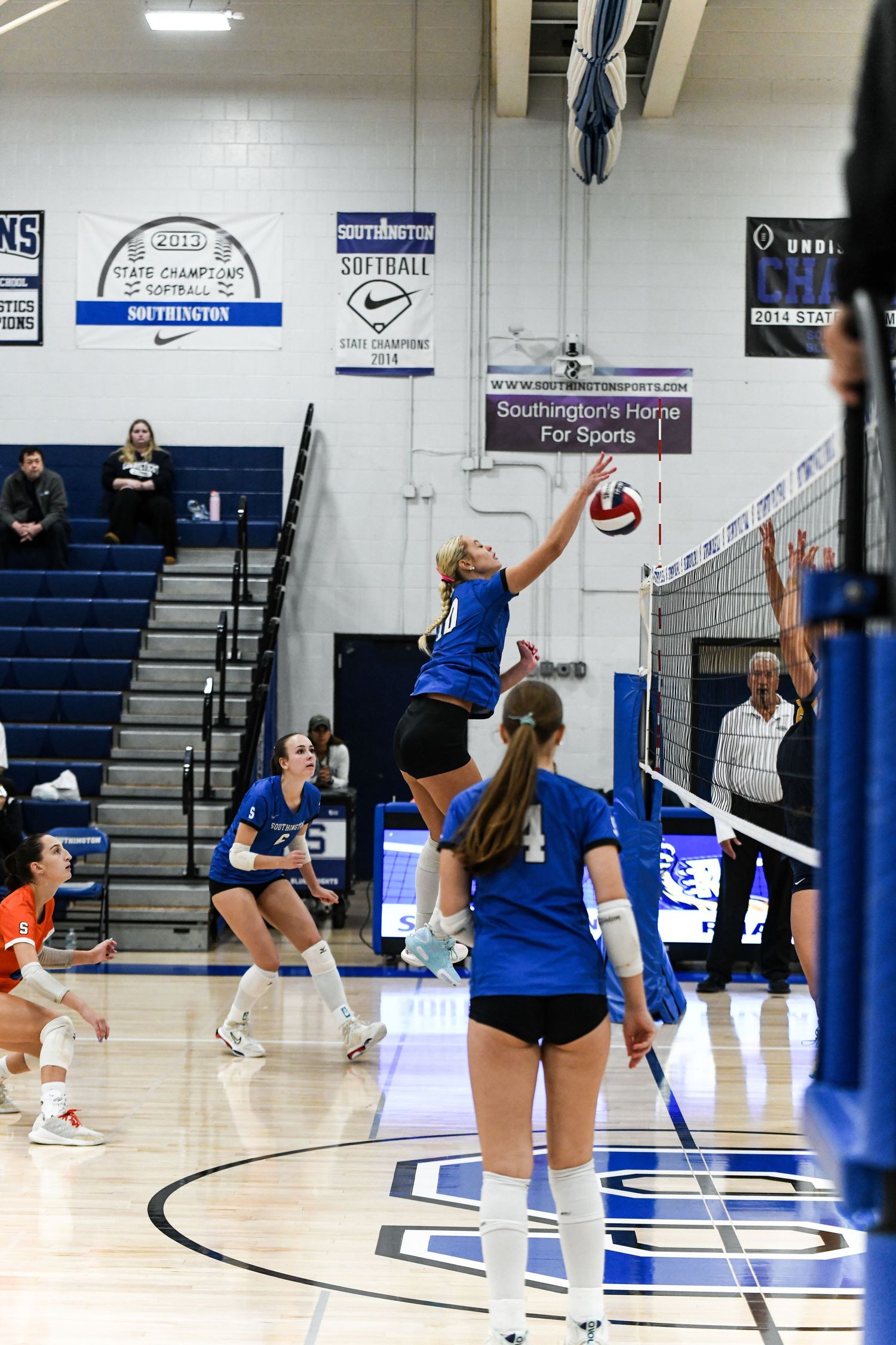 Female volleyball player jumps to spike the ball over the net during an indoor high school volleyball game.