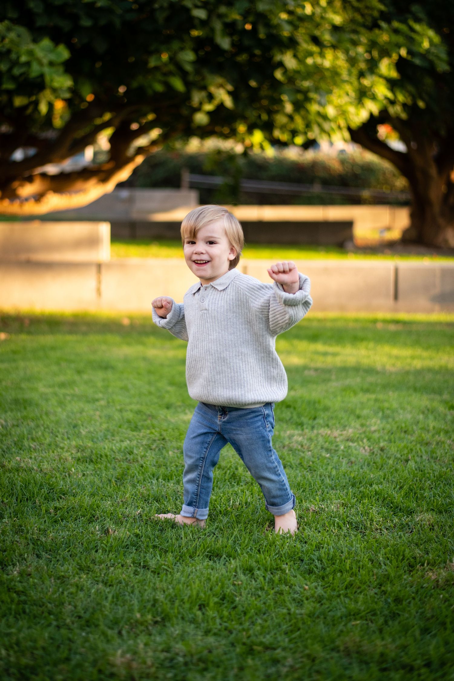 Toddler in gray sweater and jeans dancing on green grass.