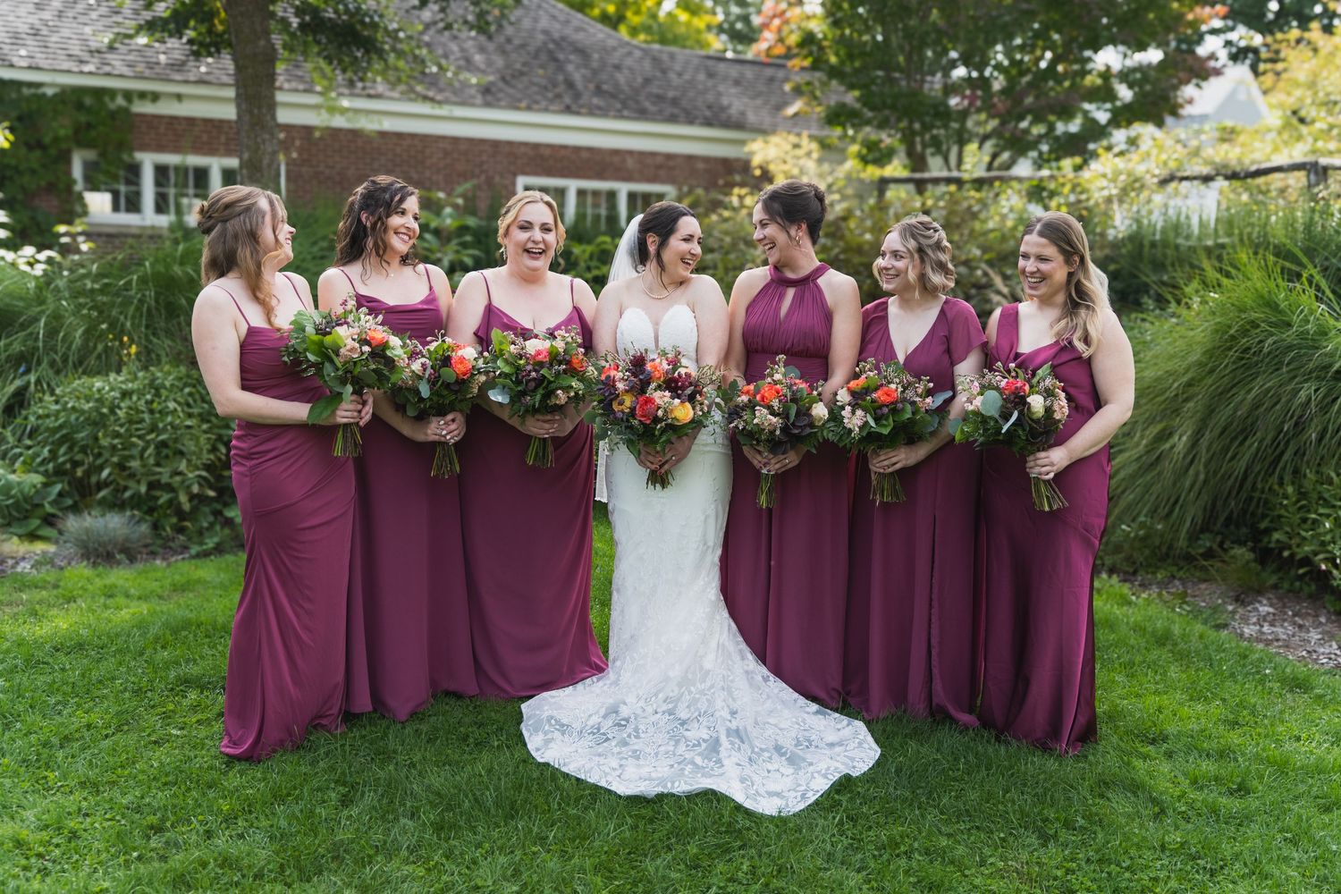 Wedding party wearing burgundy dresses poses with flowers in a garden setting.