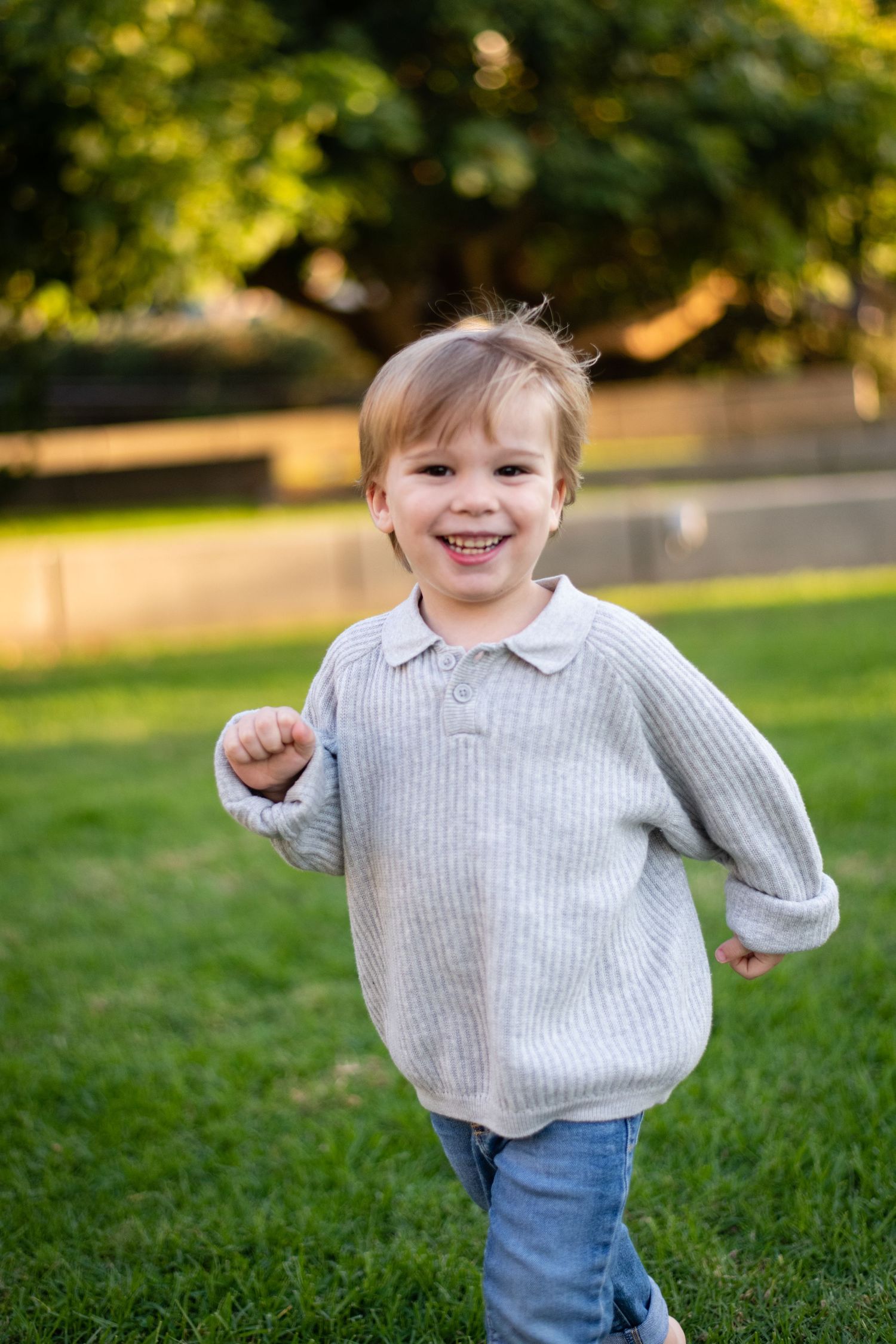 Joyful toddler playing and running on grass in a grey sweater and jeans on a sunny day.