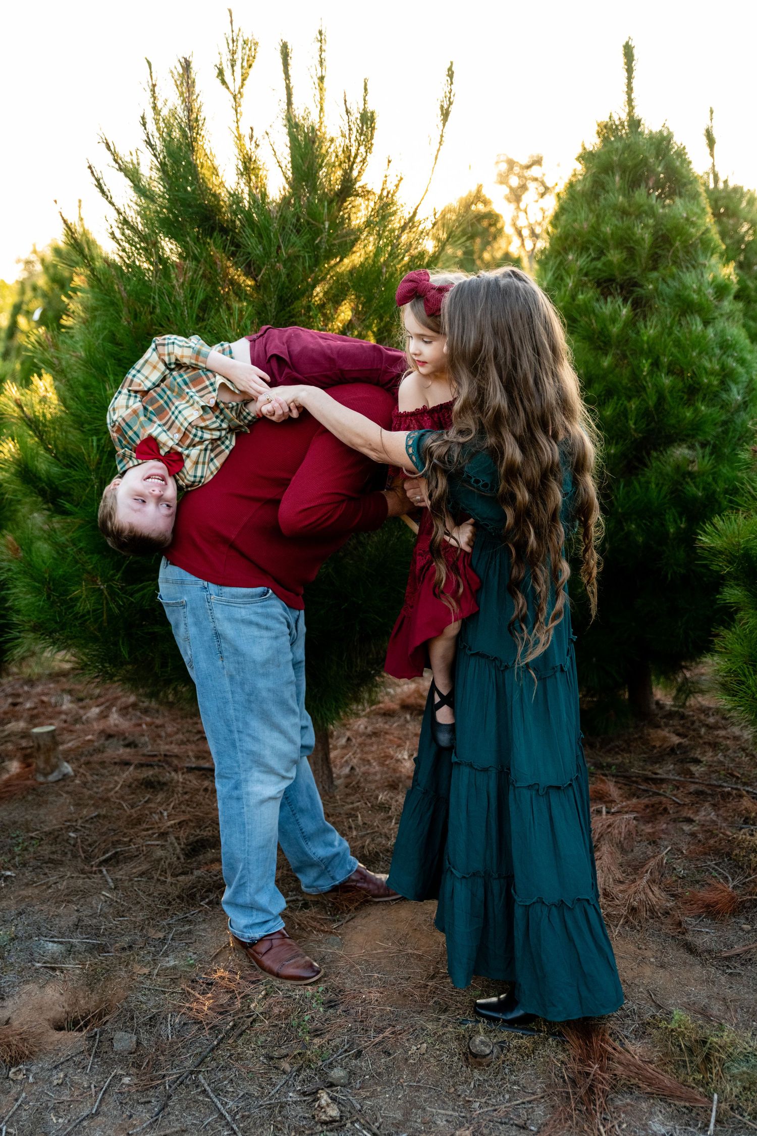 Family shares playful moment among Christmas trees at sunset.
