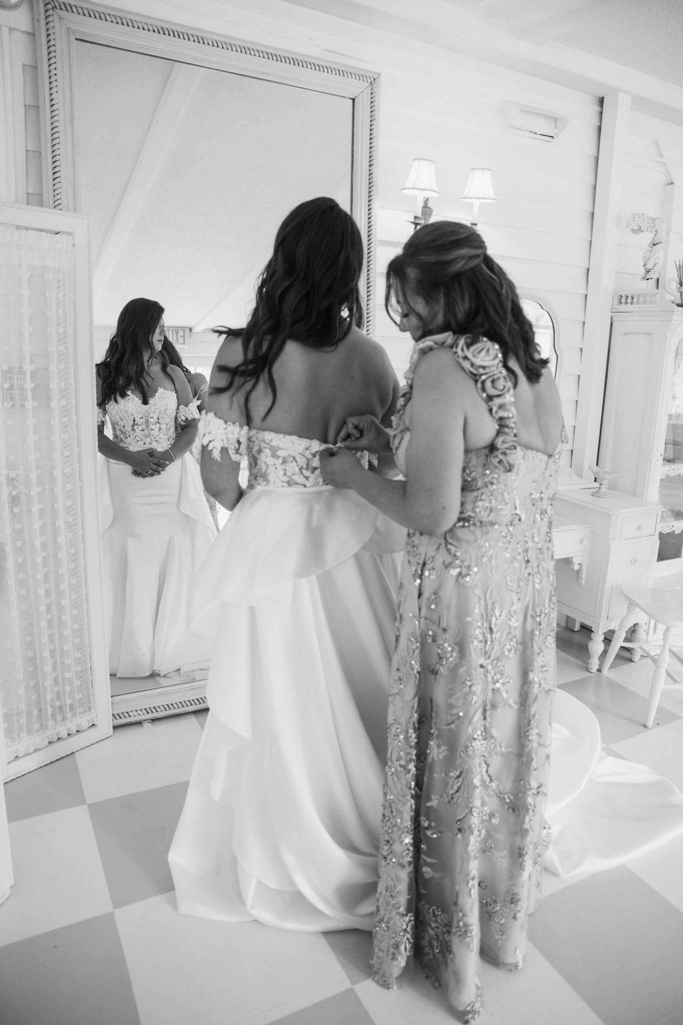 Black and white photo of bridesmaids helping with wedding dress preparations in a bright room.