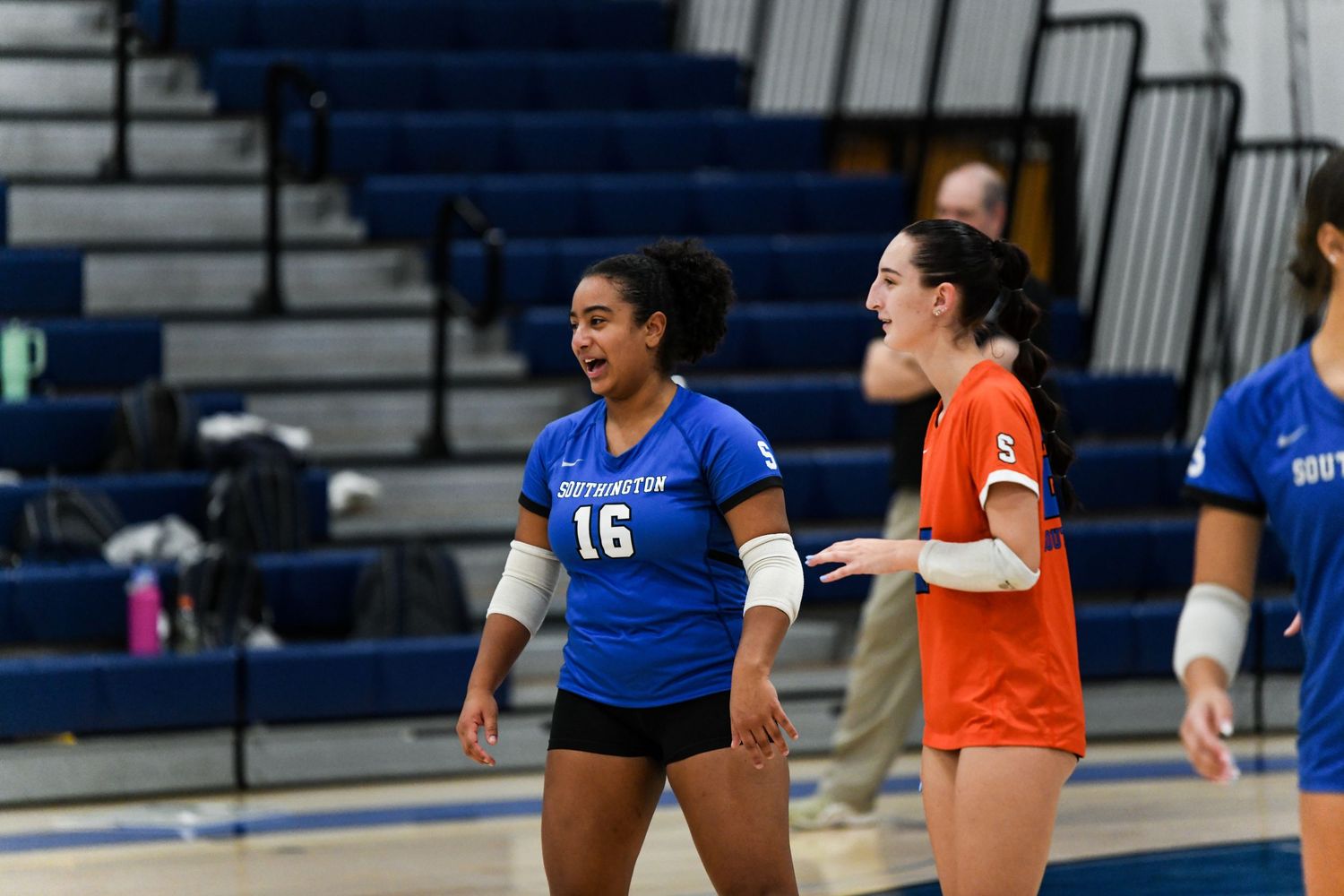 Two volleyball players in blue and orange uniforms share a laugh during a college volleyball match indoors.