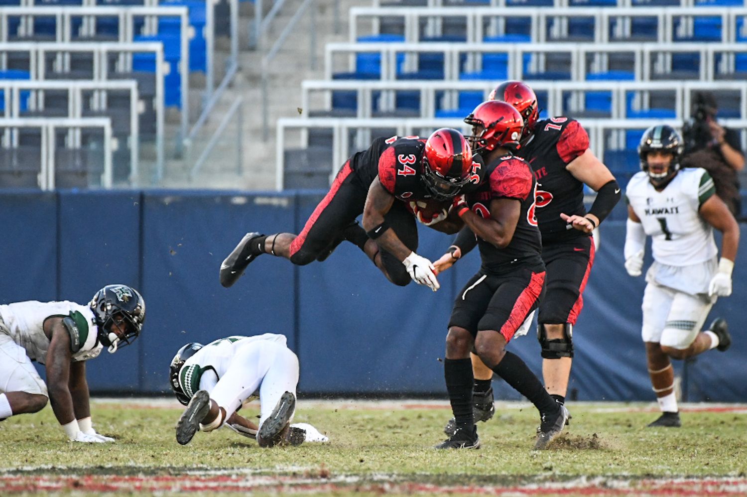 College football players in red uniforms tackle opponent during daytime game at blue stadium.