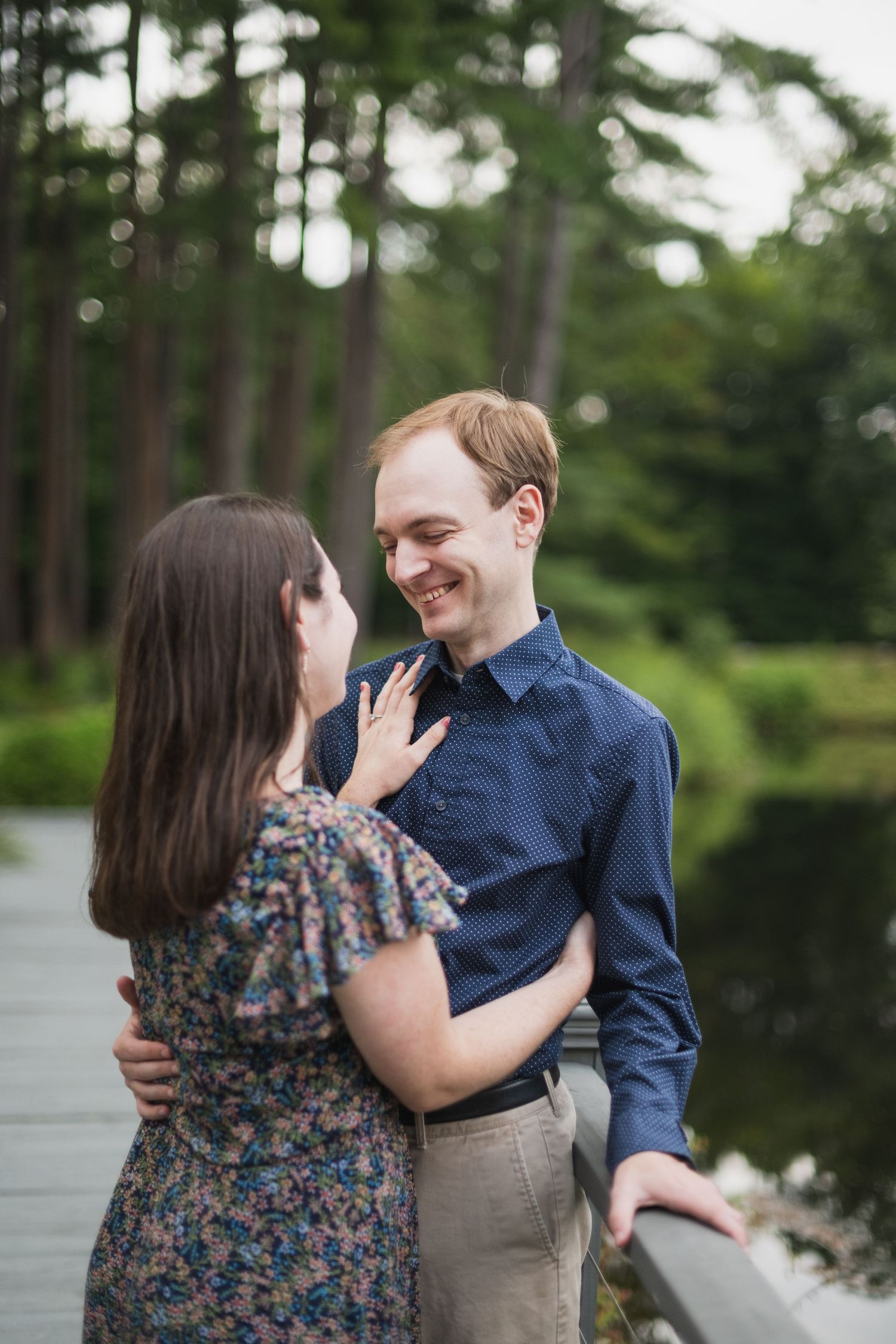 A romantic couple embraces on a wooden deck surrounded by pine trees near a lake.