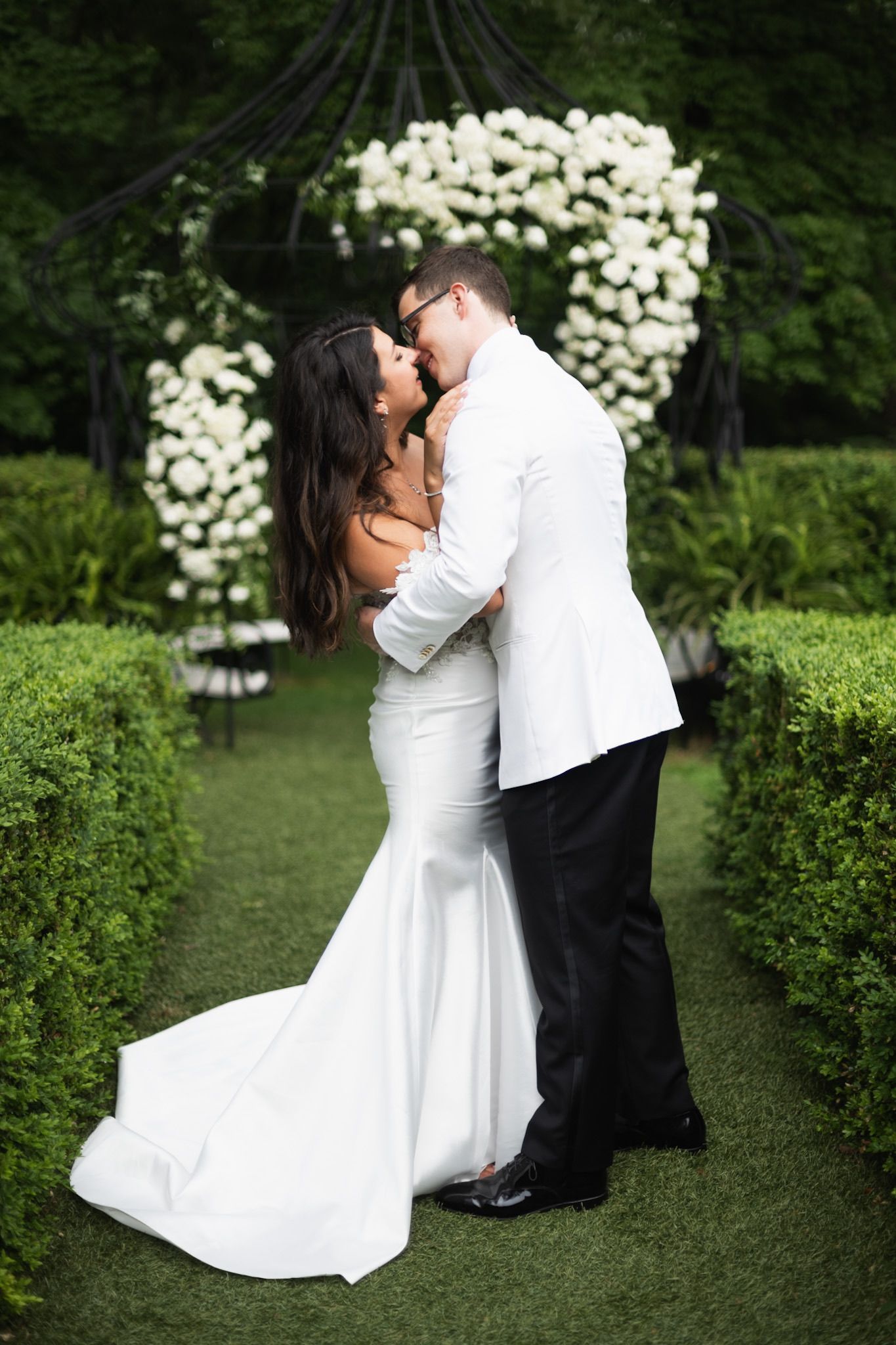 Wedding couple portraits in garden setting with white floral backdrop and manicured hedges.