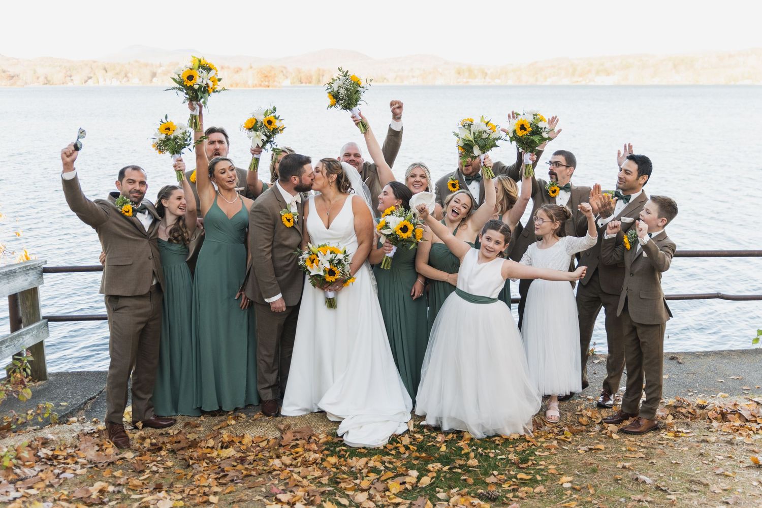 Wedding party celebrates on a lakeside beach in autumn with green and brown attire.