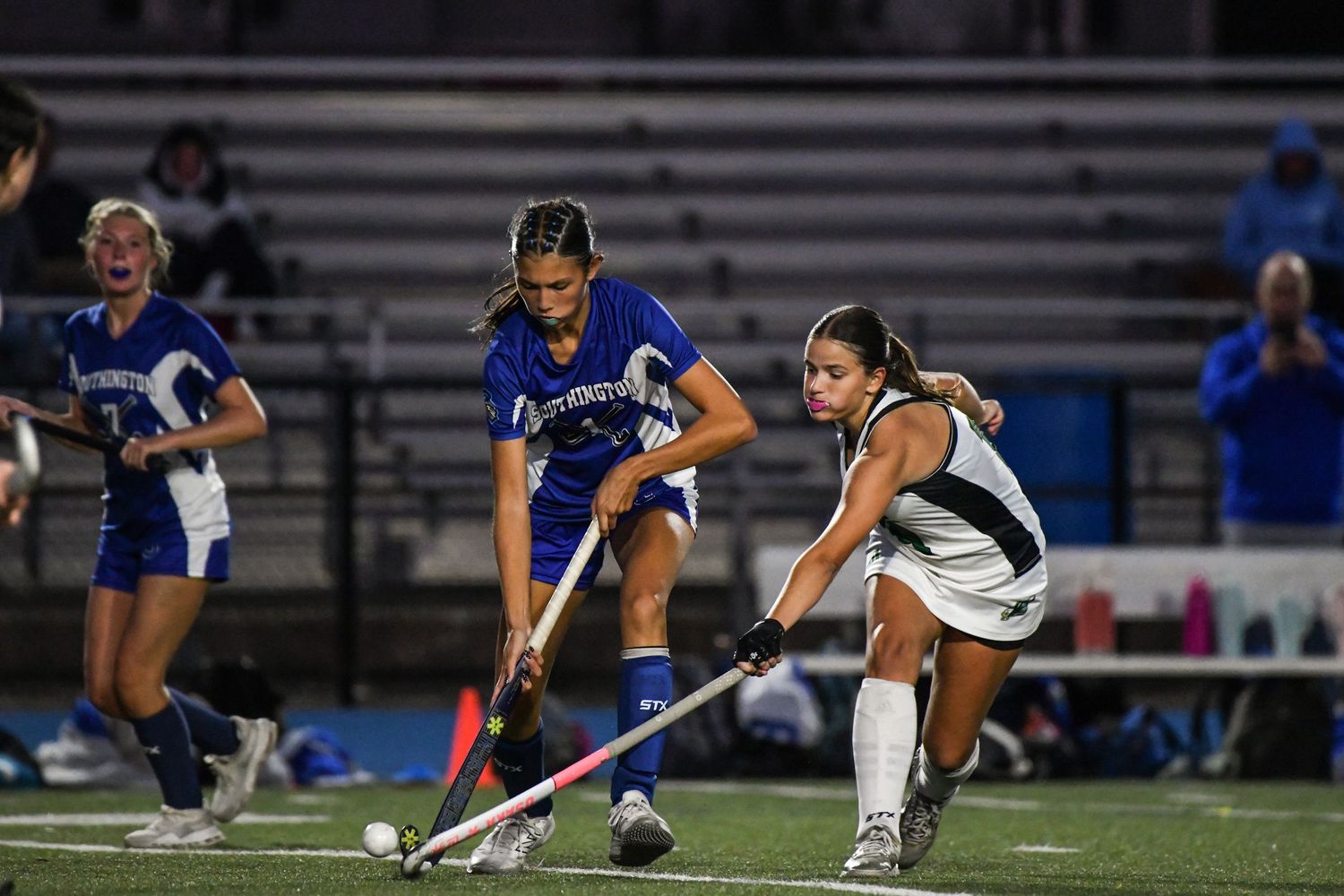 Two field hockey players compete for the ball during a nighttime high school field hockey game.