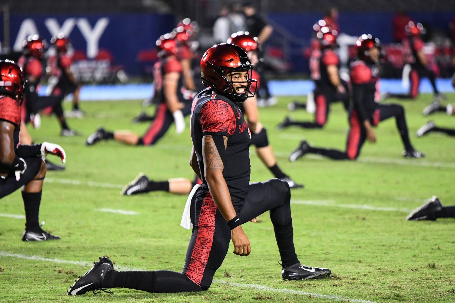 Football players in red and black uniforms stretching on the field during a night game warm-up session.