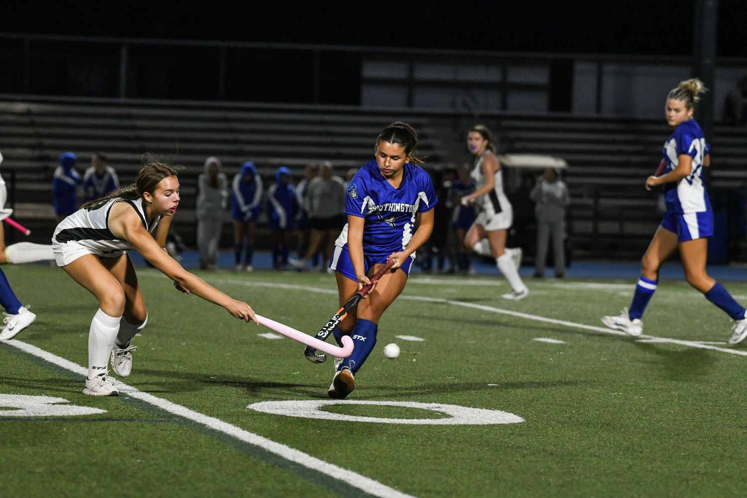 Two female field hockey players compete for the ball during a night game on a turf field.