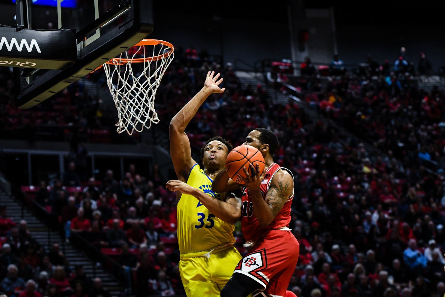 Basketball player in yellow jersey attempts to block a shot near the hoop during an indoor college basketball game.