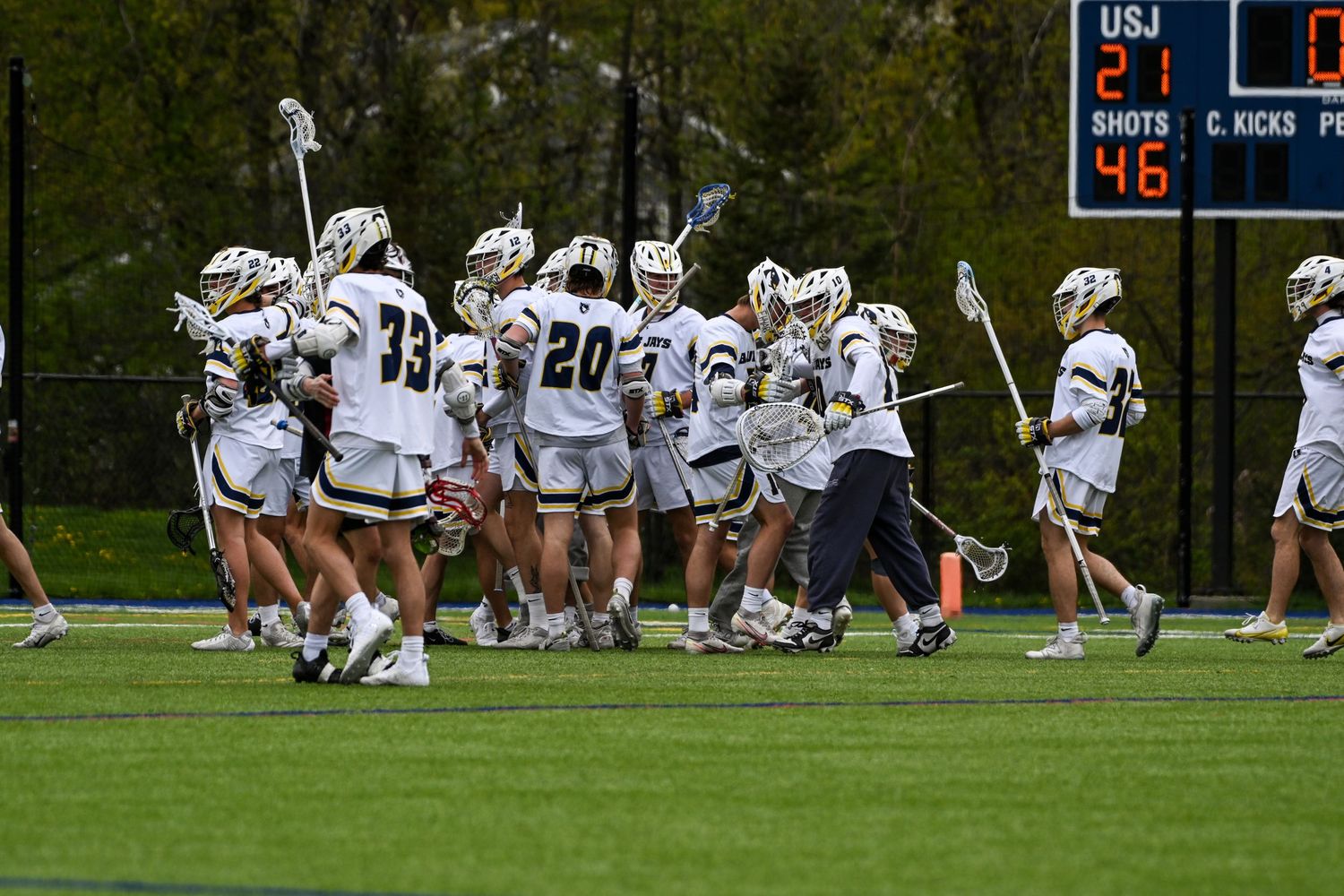 Lacrosse team players in white and gold uniforms celebrating on field with scoreboard in background.