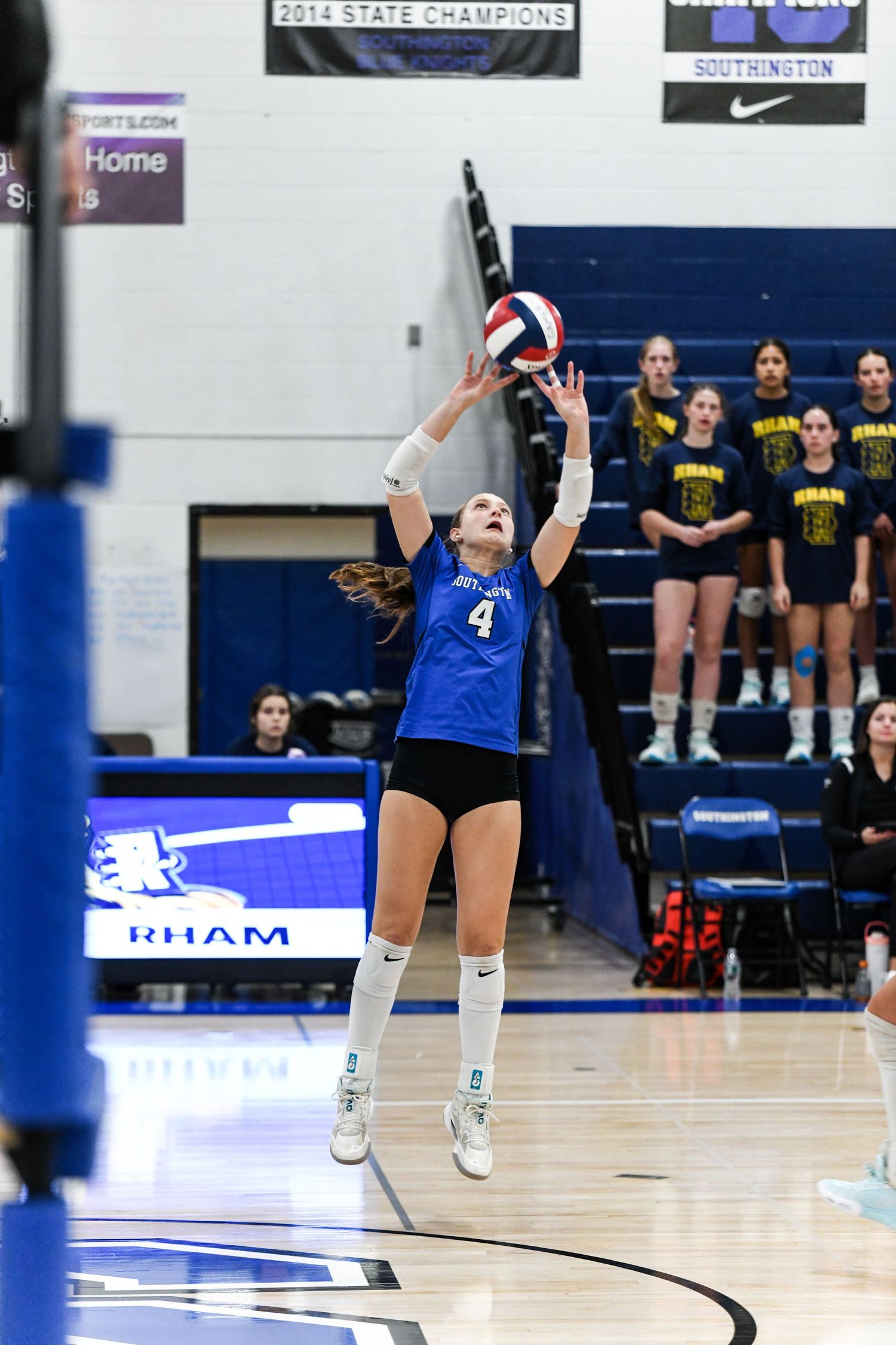 Player #4 in blue uniform sets a volleyball during a high school gym match, with teammates watching from the sideline.