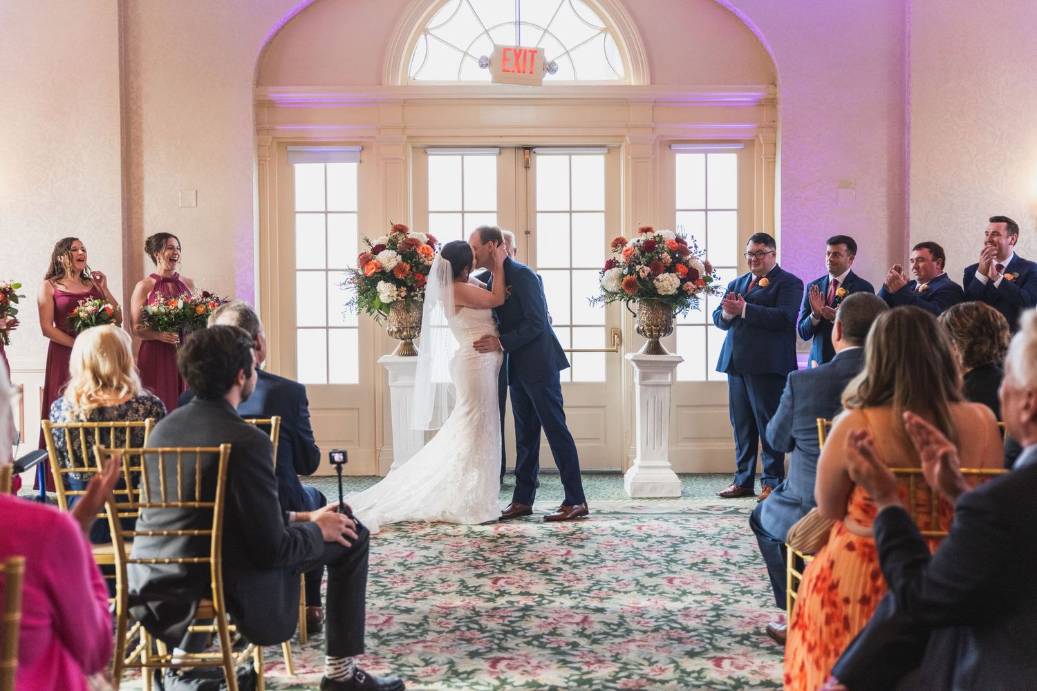 Wedding guests mingle and dance in an elegant ballroom with arched windows and soft purple lighting.