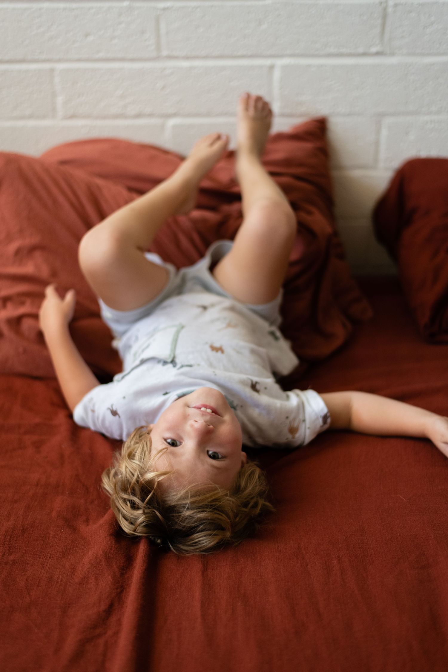 Child lying playfully on rust-colored bedding against white brick wall background.